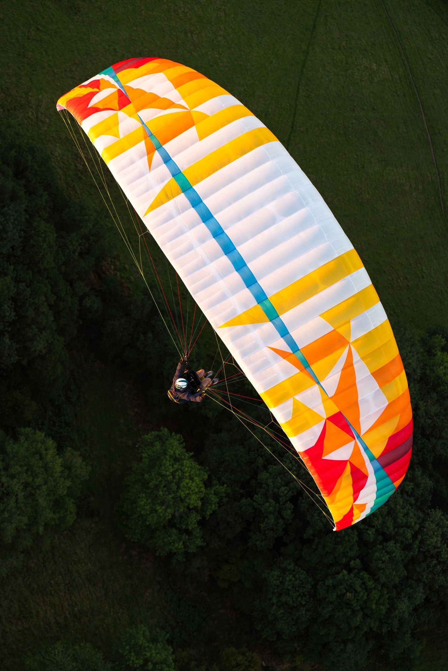A colorful paraglider with yellow, orange and red geometric patterns soars above green forest canopy at sunset.