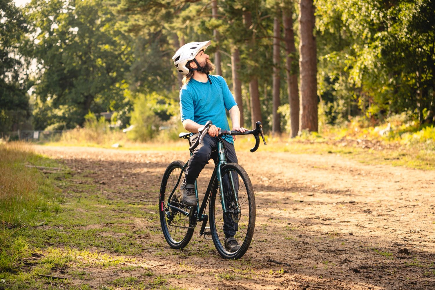 Cyclist in bright blue jersey and white helmet stands with road bike on dirt trail surrounded by trees in autumn.