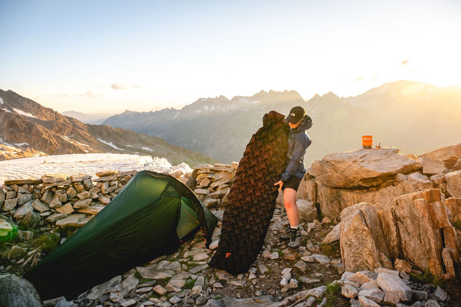 A camping tent sits perched on a rocky mountain ridge at sunset with someone viewing the majestic alpine landscape.