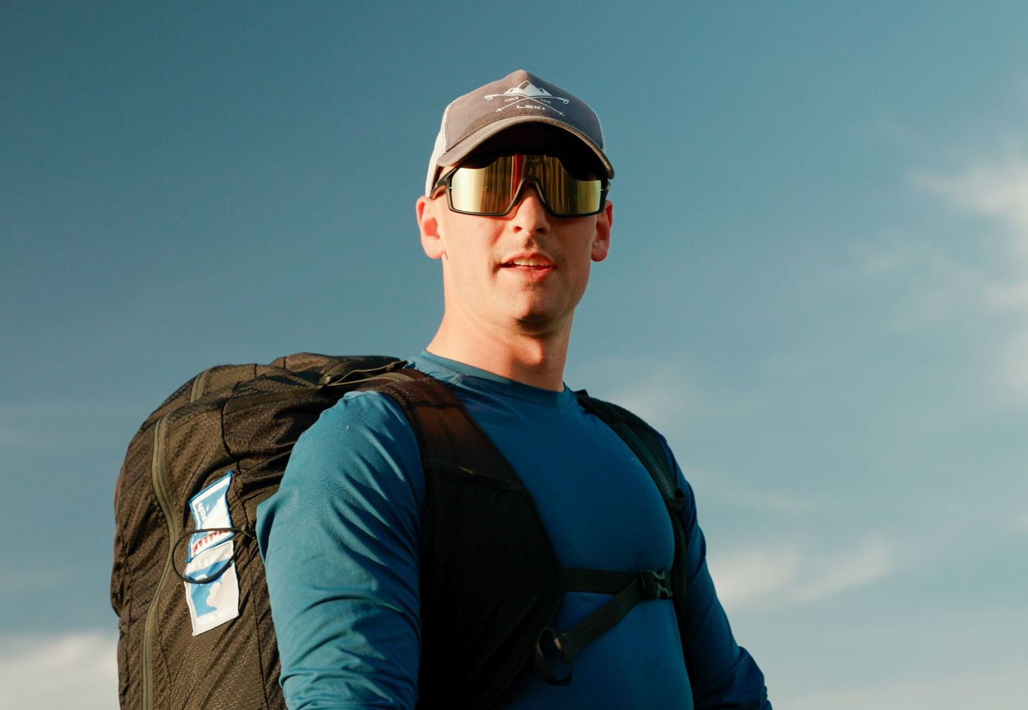 Hiker wearing sunglasses and a baseball cap stands against blue sky with a backpack on a sunny day.