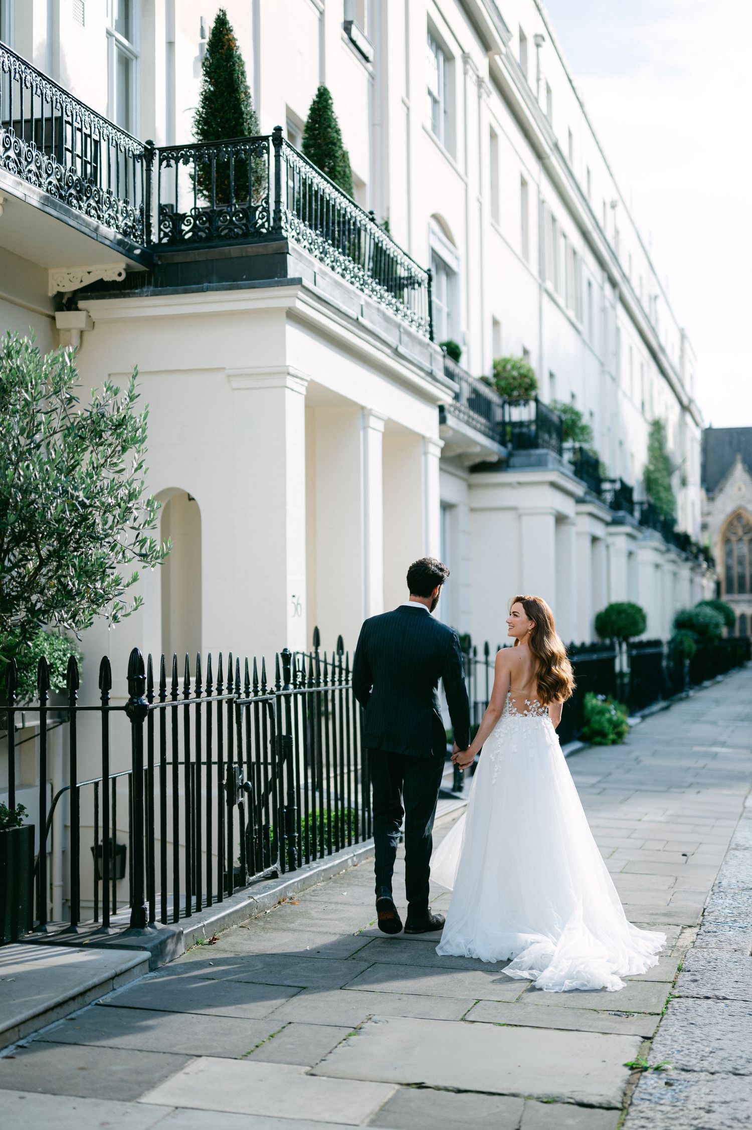 Newlyweds walking along elegant white townhouses with black iron railings and manicured topiaries.