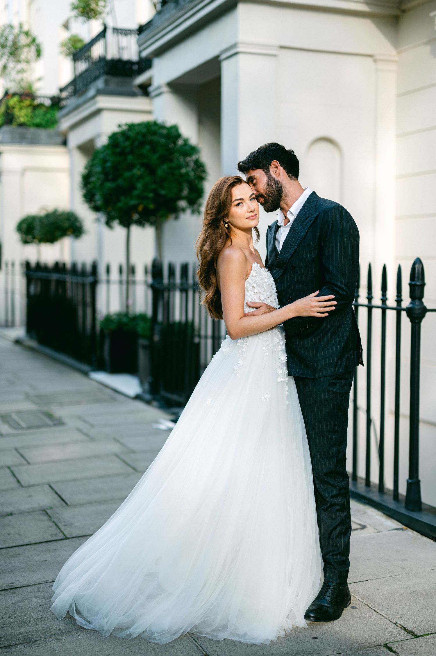 A romantic wedding couple poses together on an elegant city street with white buildings and iron fencing.