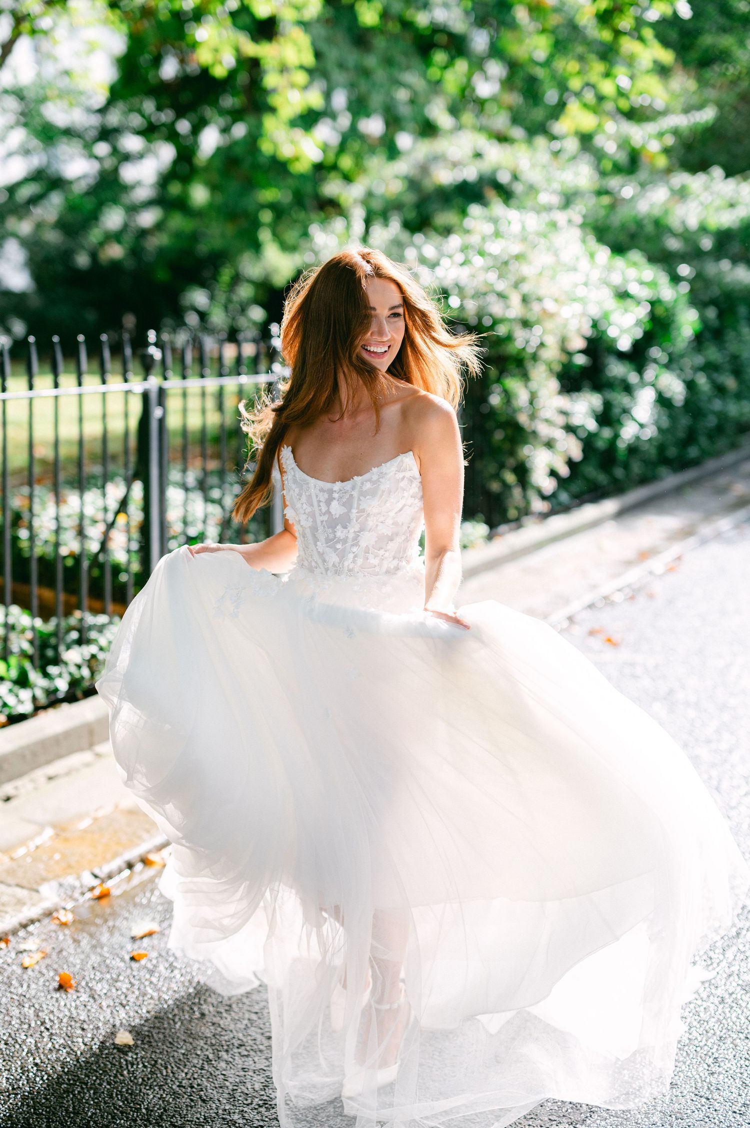 A joyful person in a flowing white wedding dress twirls on a path surrounded by lush green foliage and white flowers.