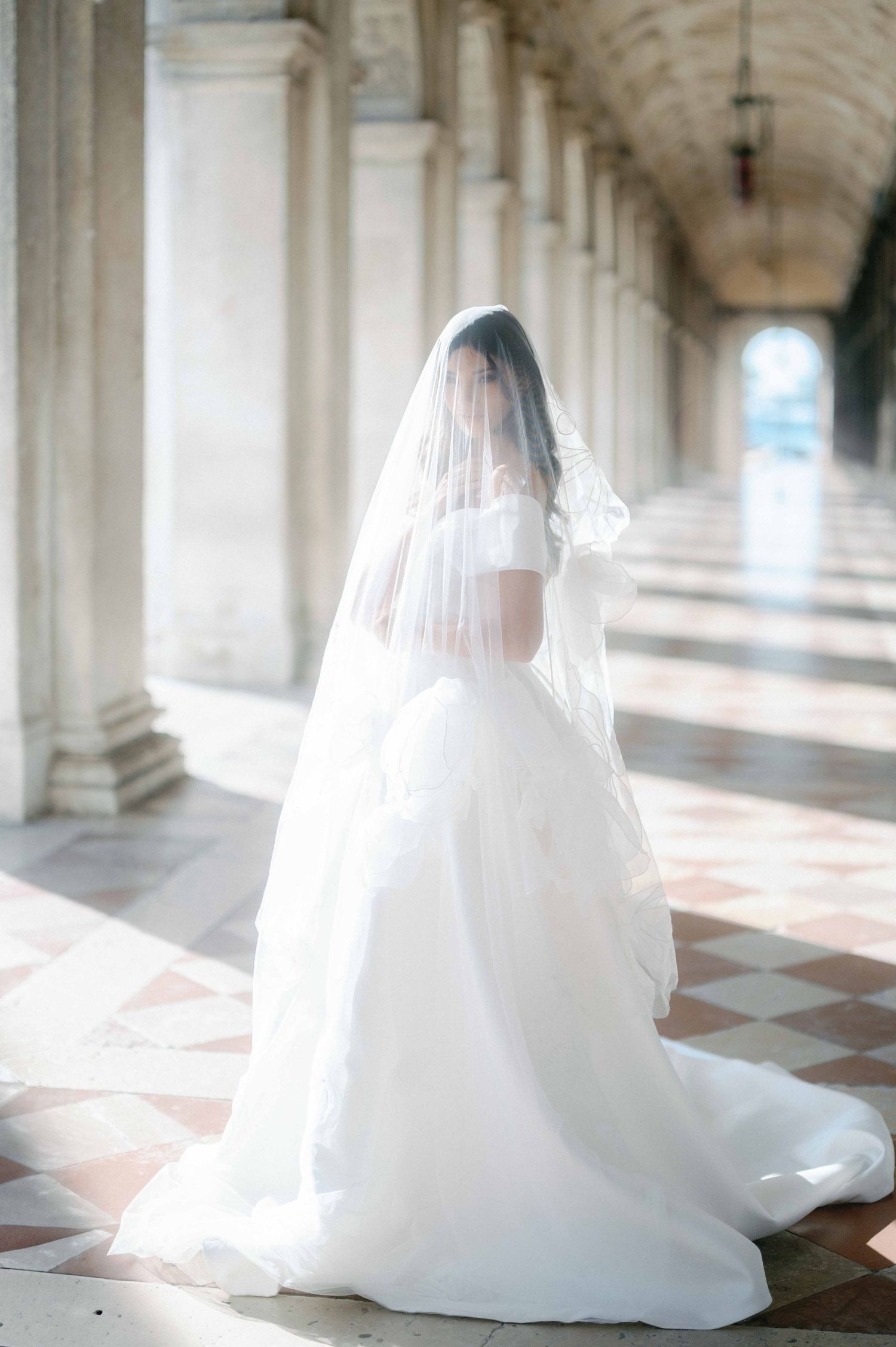 Bride in flowing white wedding gown and veil stands in elegant colonnade with checkered marble floor and arched ceiling.