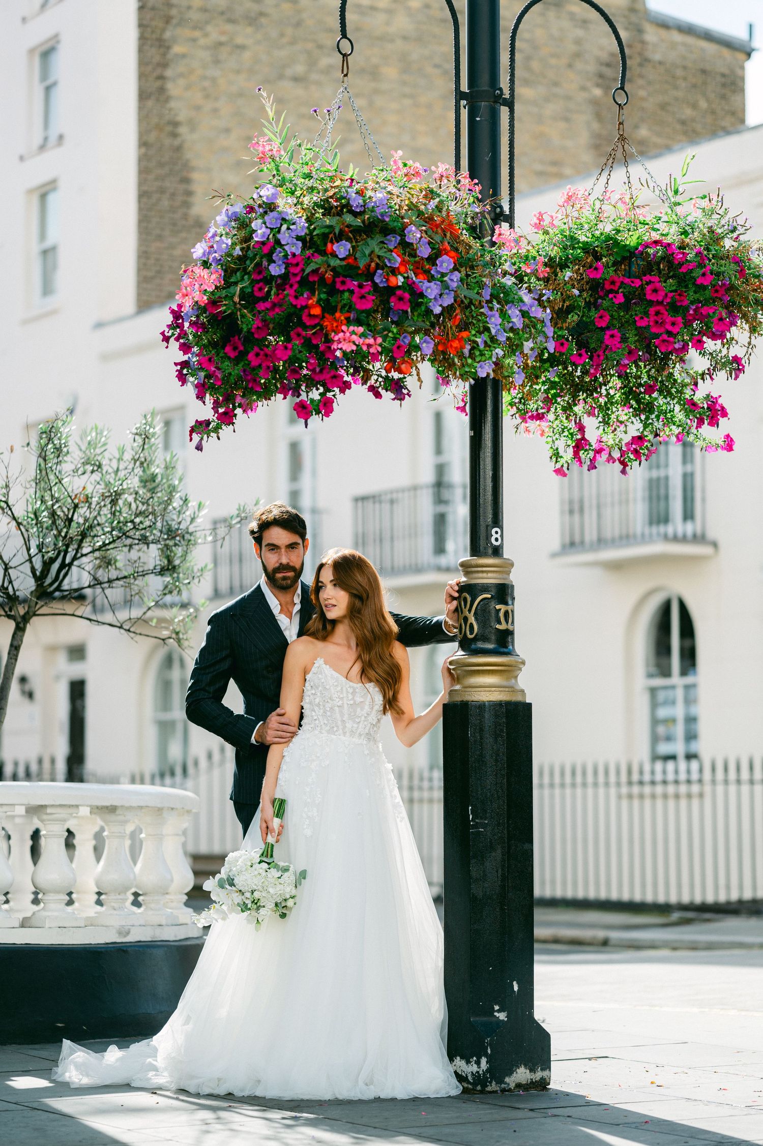 A couple poses near an ornate lamppost with vibrant hanging flower baskets on a city street with white buildings.