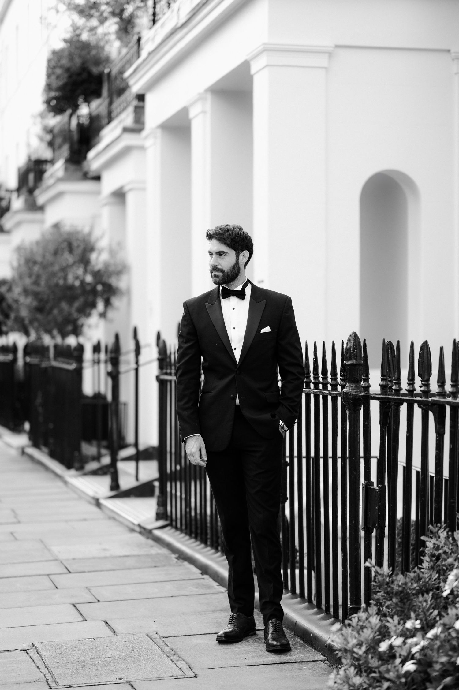 Black and white portrait of a man in a tuxedo and bow tie standing elegantly by a white townhouse with iron railings.