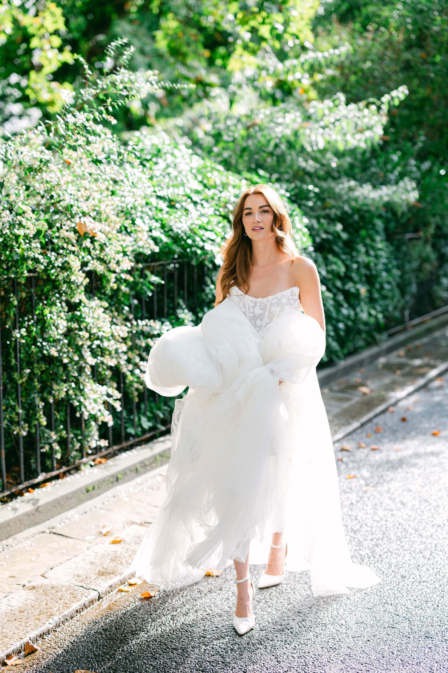 A bride in a flowing white wedding dress runs joyfully down a garden path surrounded by blooming white flowers.