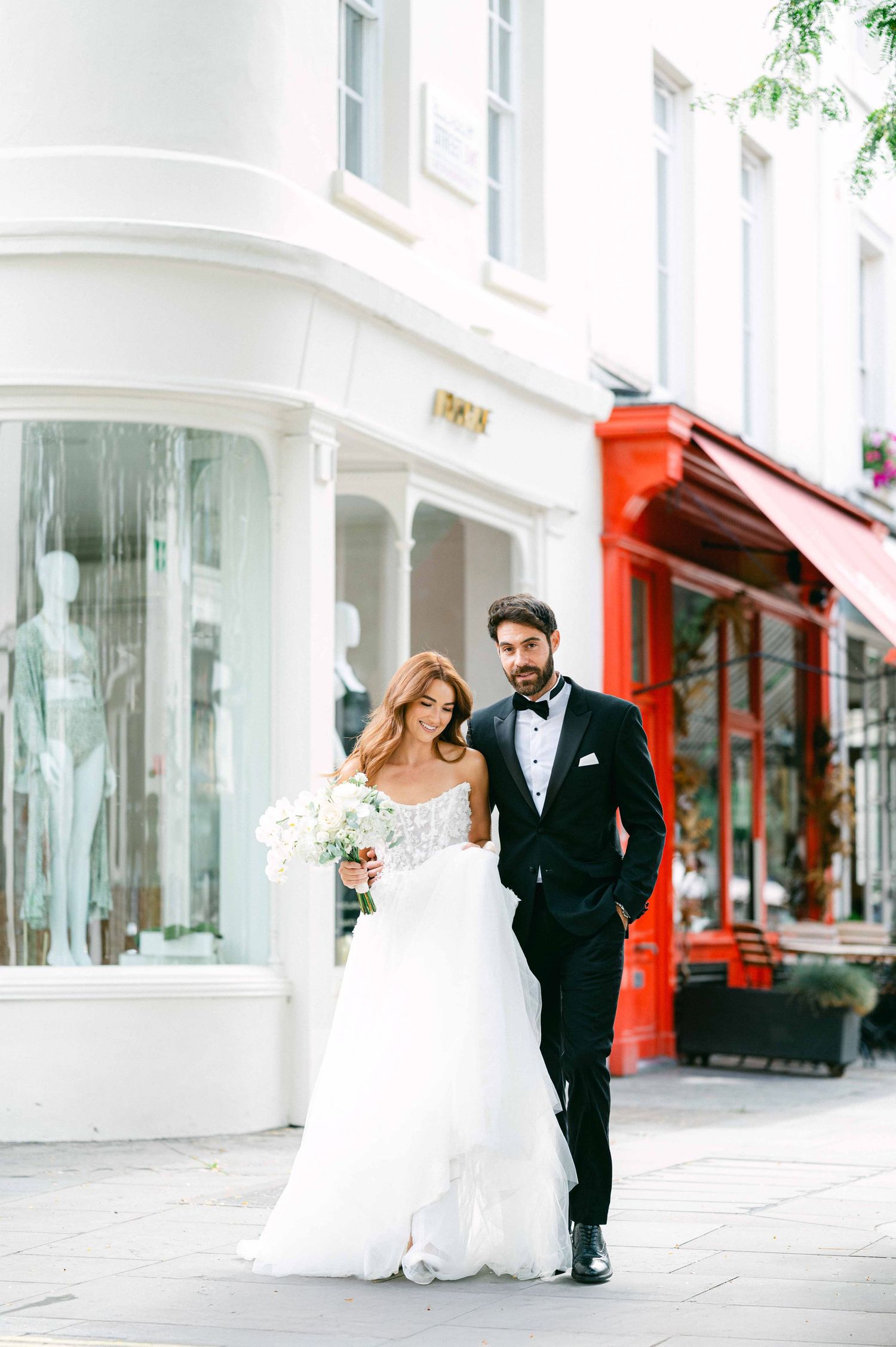 A couple in wedding attire walking together on a charming city street with white buildings and red storefront.