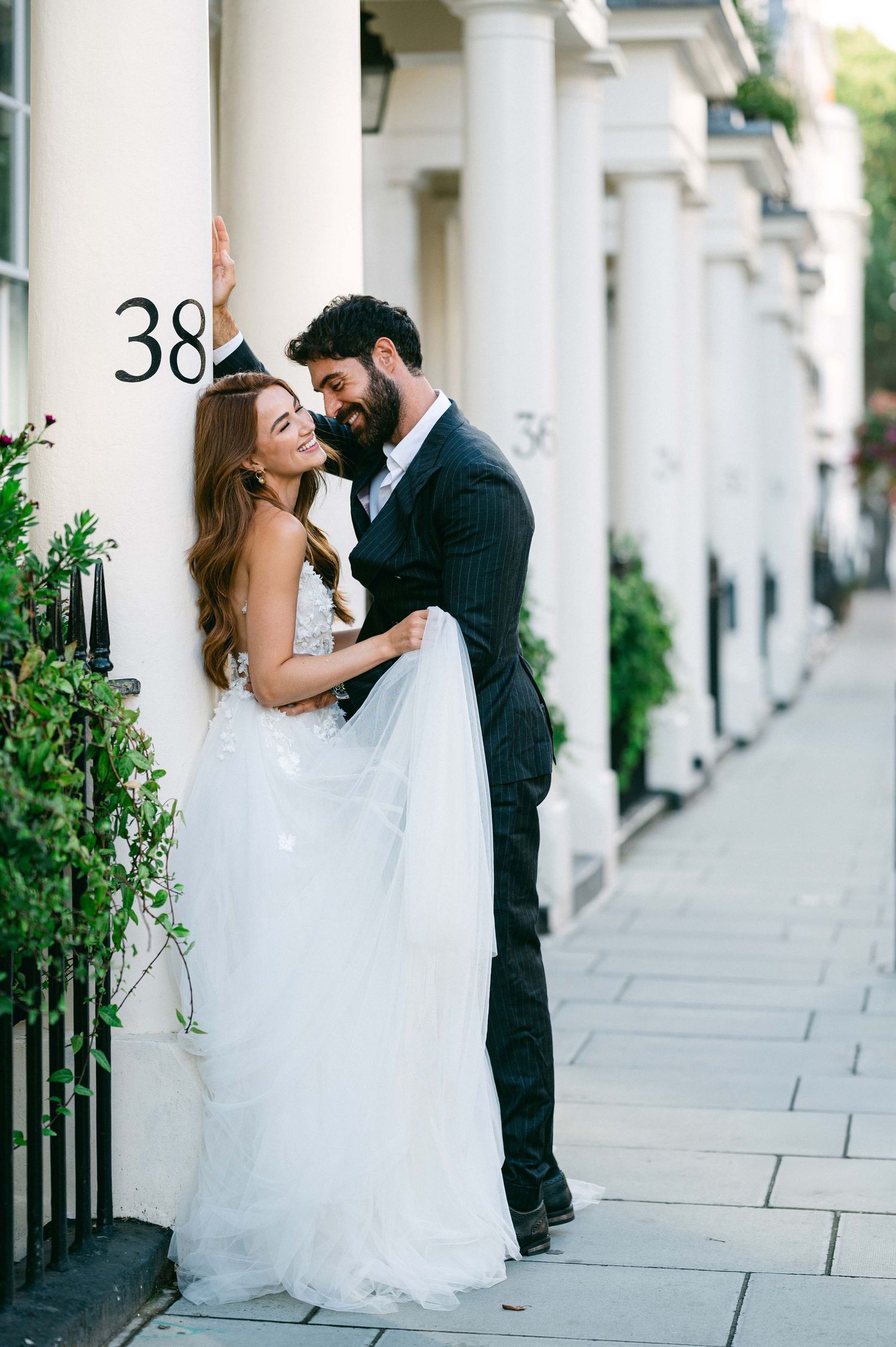 A couple in wedding attire embraces on a charming London street with white columns and house number 38.