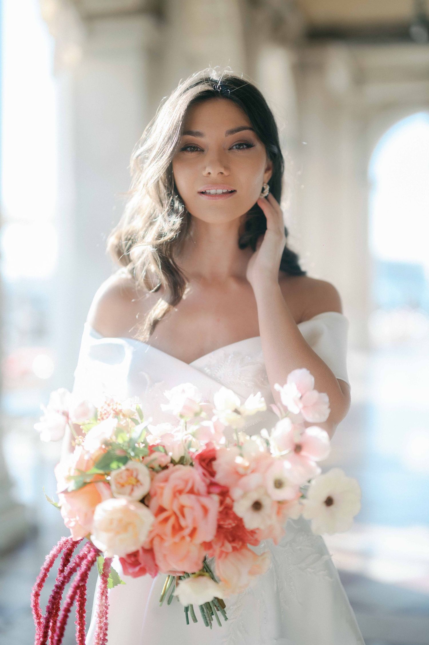 A bride in an off-shoulder white wedding dress holds a beautiful bouquet of pink and peach flowers in a bright setting.