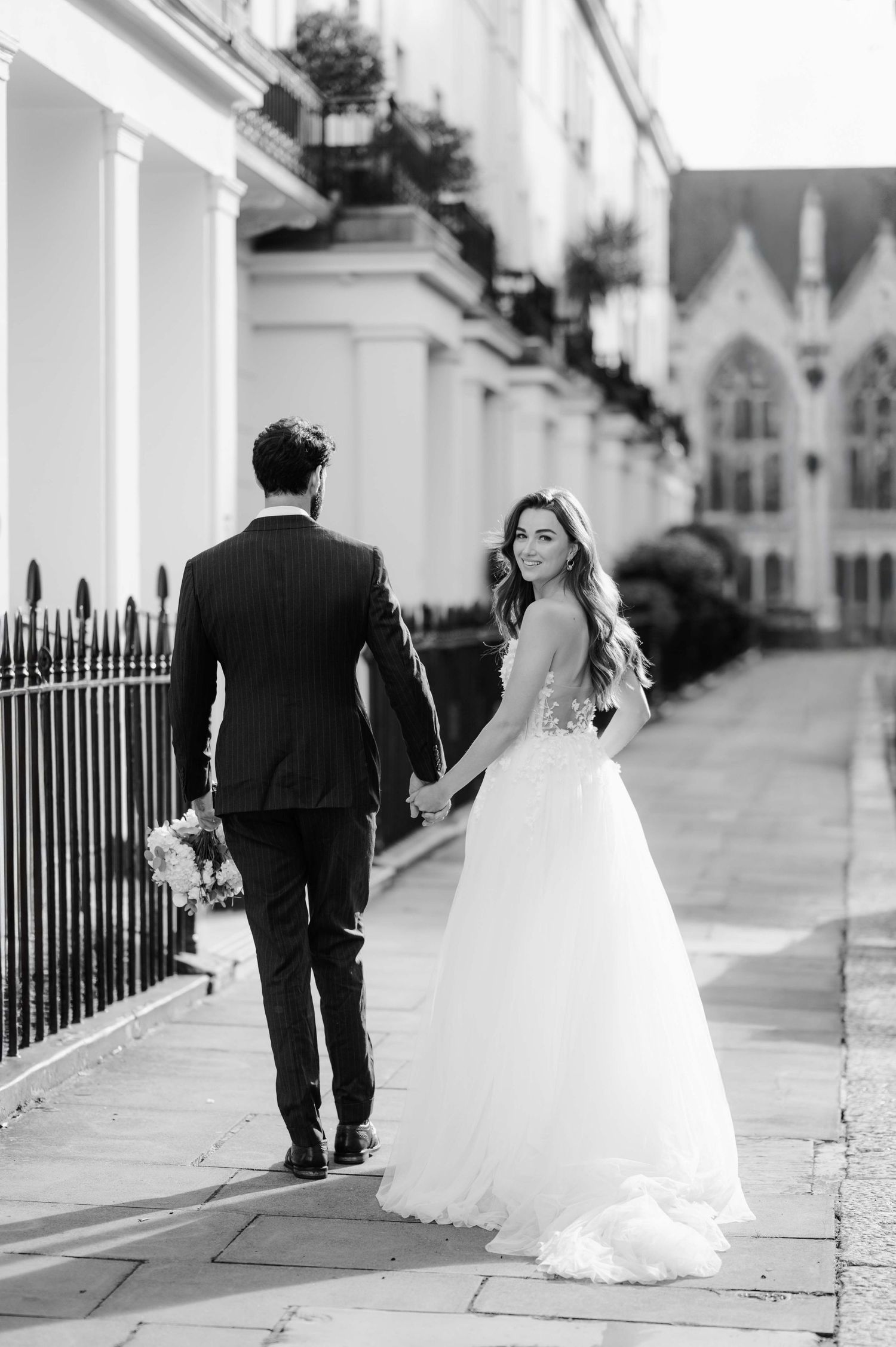 Black and white photo of a bride in a flowing gown looking back while walking with groom along an elegant street with iron railings.