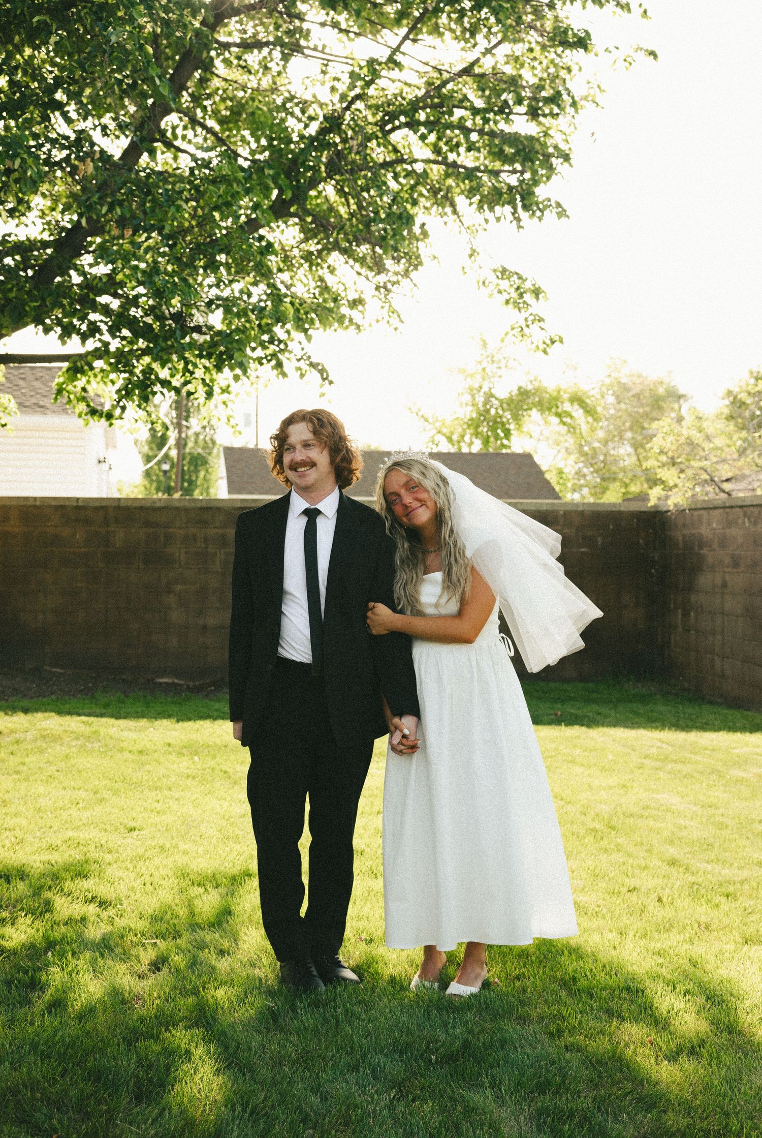 Wedding couple shares playful moment while posing outdoors in garden setting with green trees and lawn.