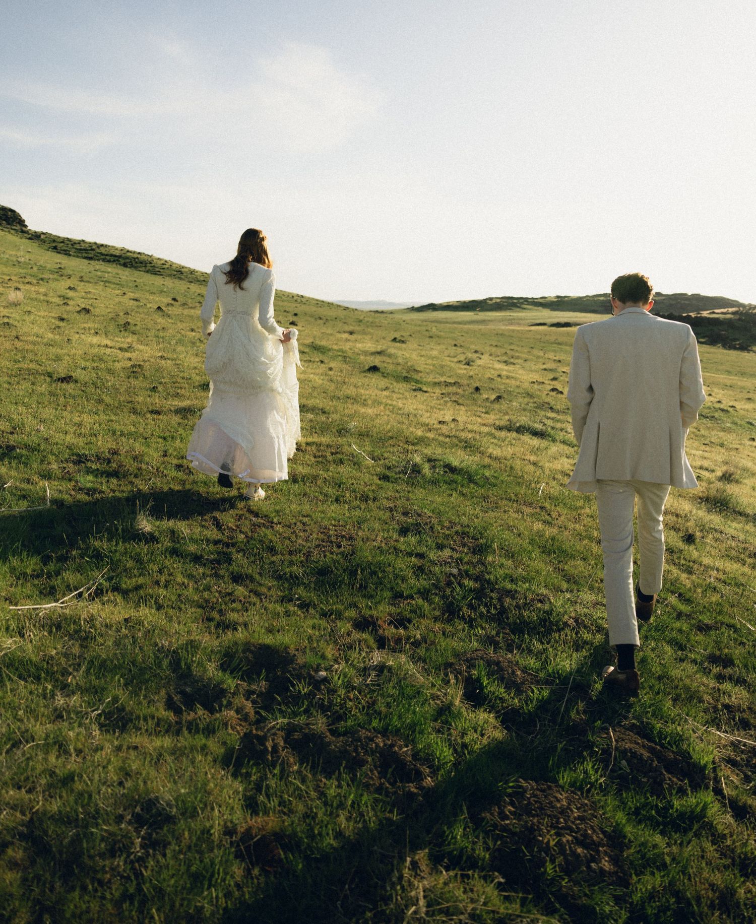 Two people in wedding attire walk up a grassy hillside on a sunny day creating long shadows behind them.