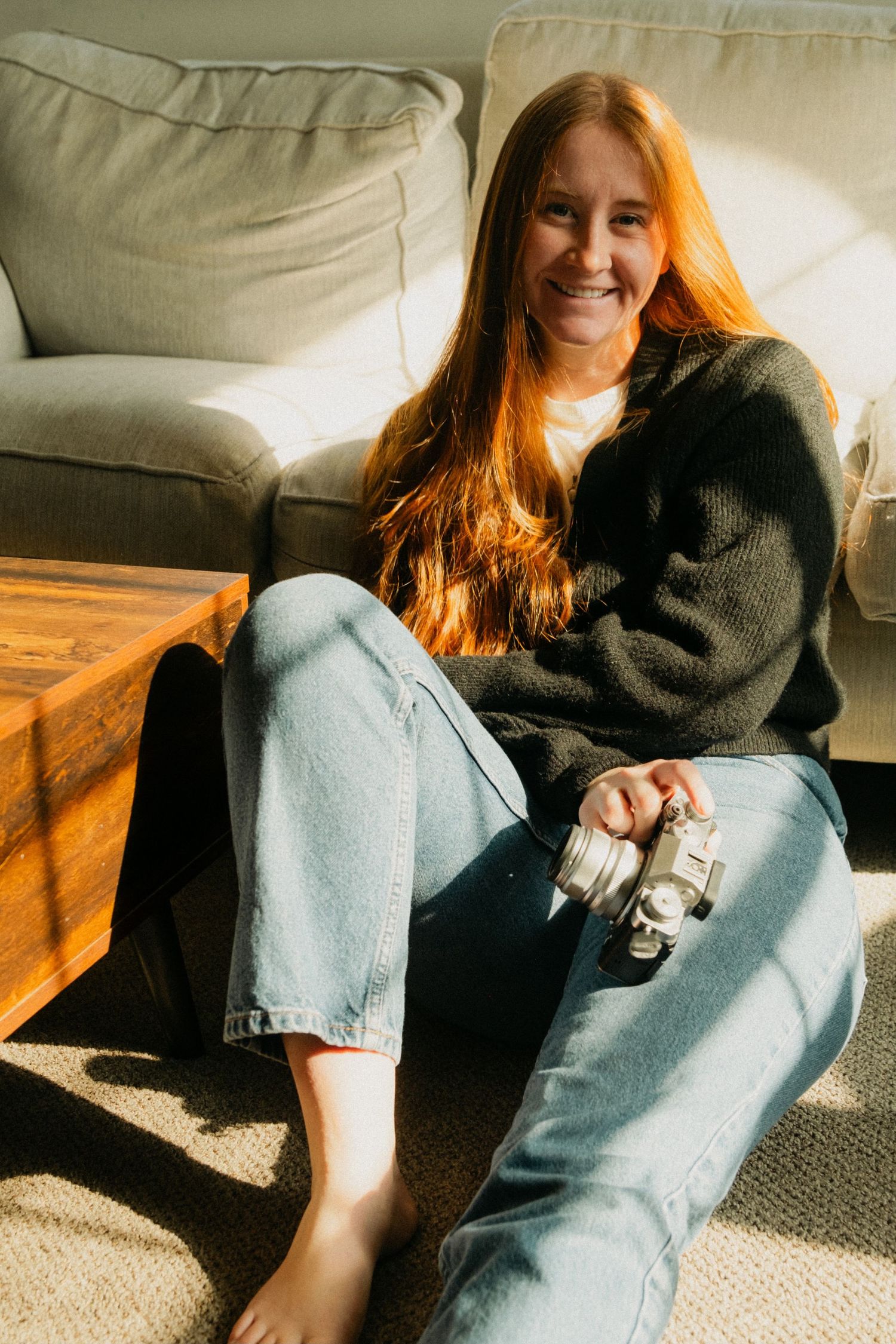 A person with long red hair relaxes on a couch in a black sweater and jeans, sitting in warm sunlight.