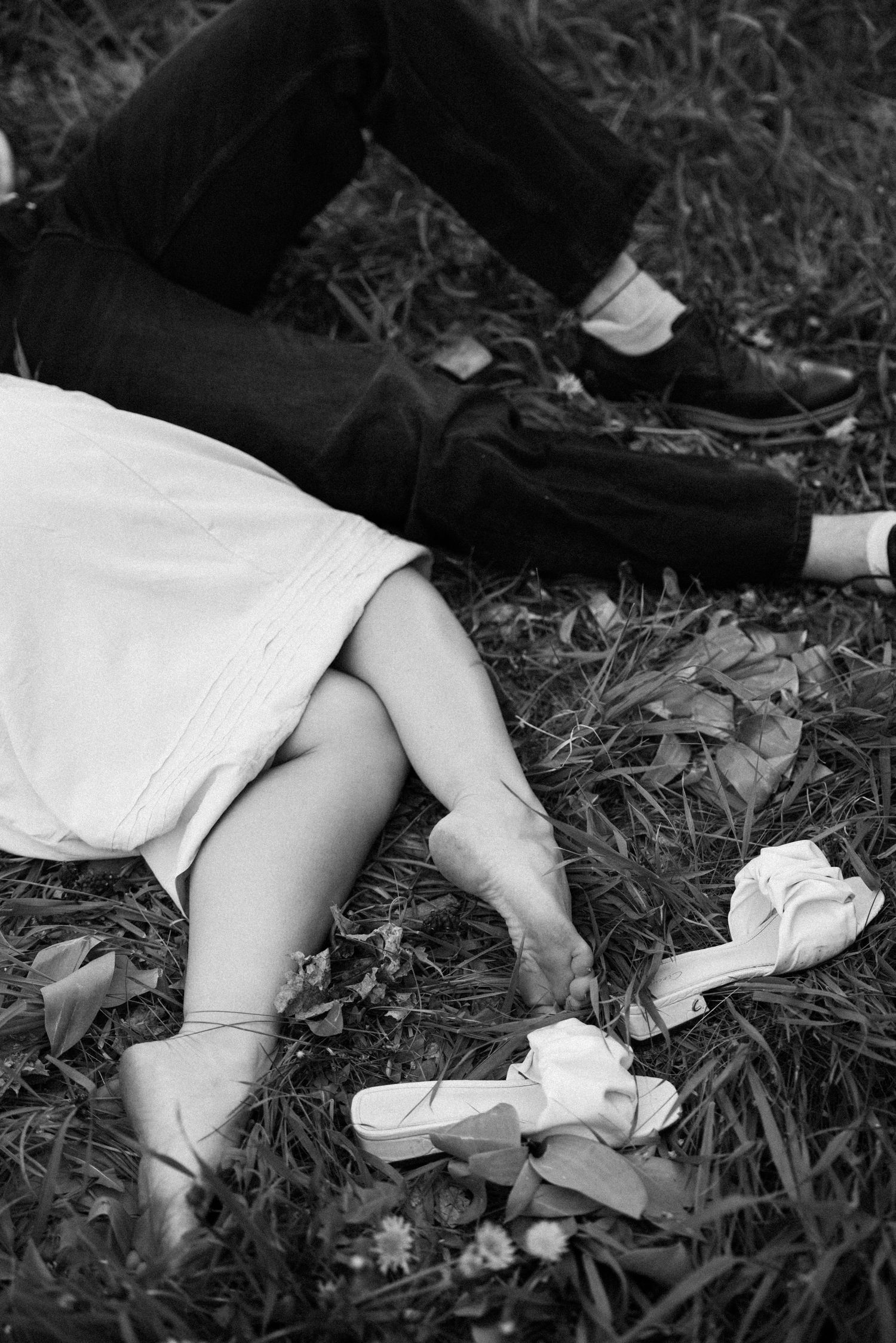 Black and white artistic photograph of two people reclining on grass, one wearing a white dress and ballet shoes.