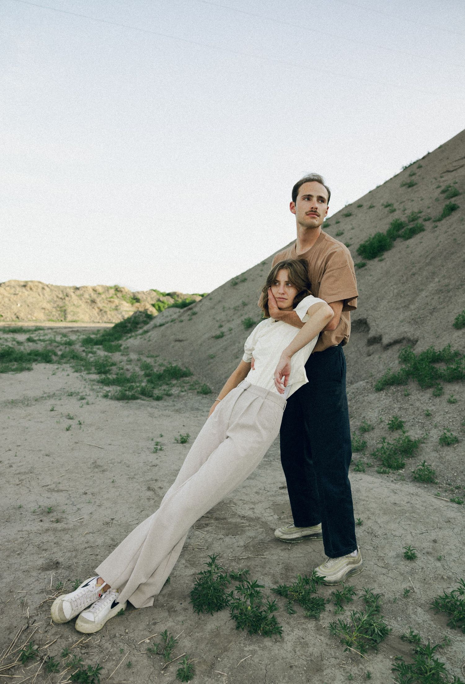 Two people pose together while leaning against each other on a desert hillside in casual earth-toned clothing.