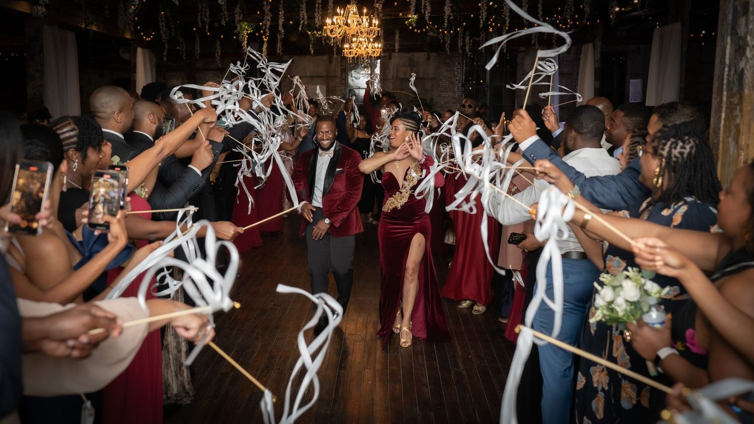 Wedding party celebrating on the dance floor with white ribbon wands under an elegant crystal chandelier.