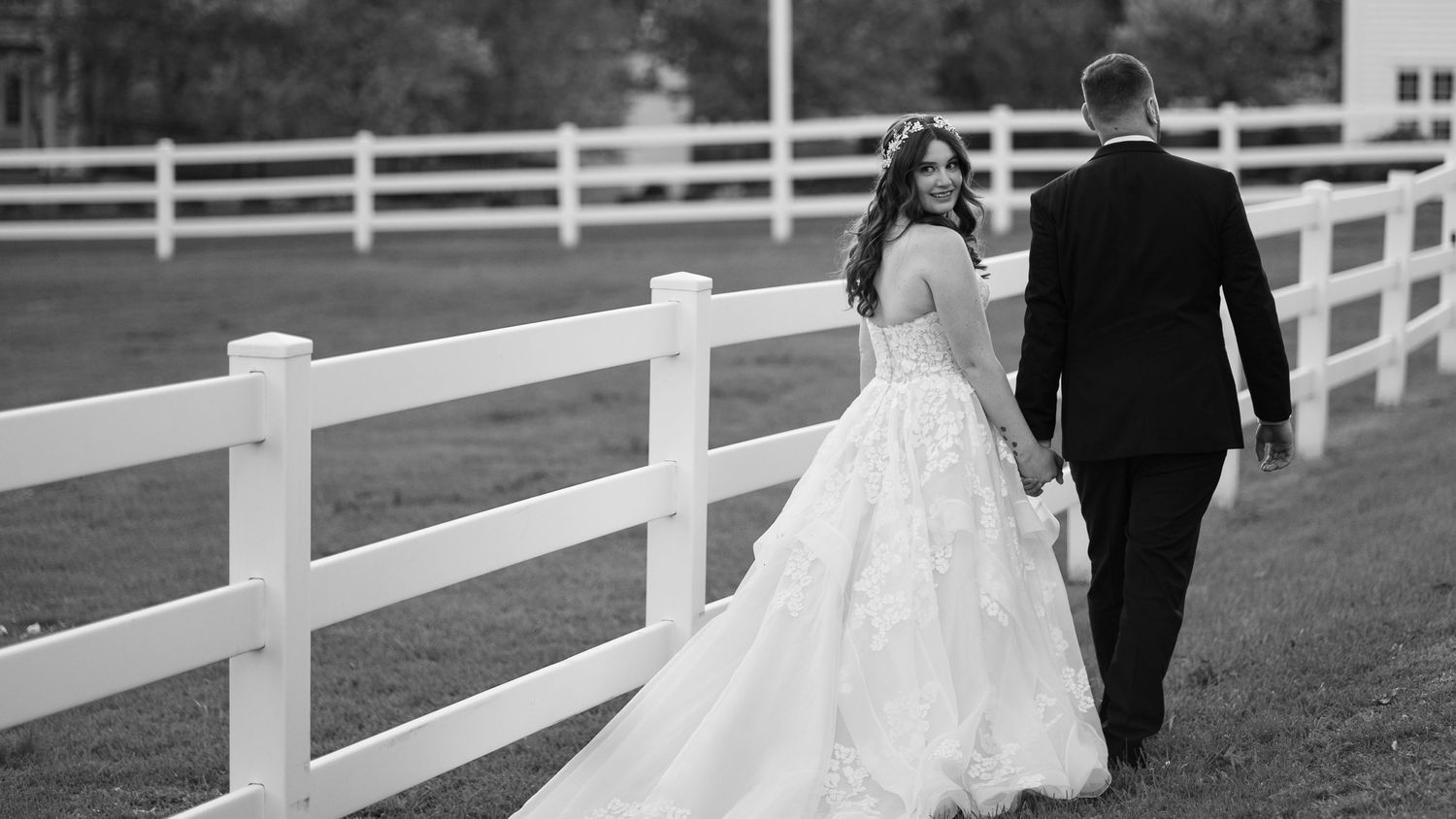 Black and white photo of a couple walking along a white fence on their wedding day, with bride wearing flowing gown.