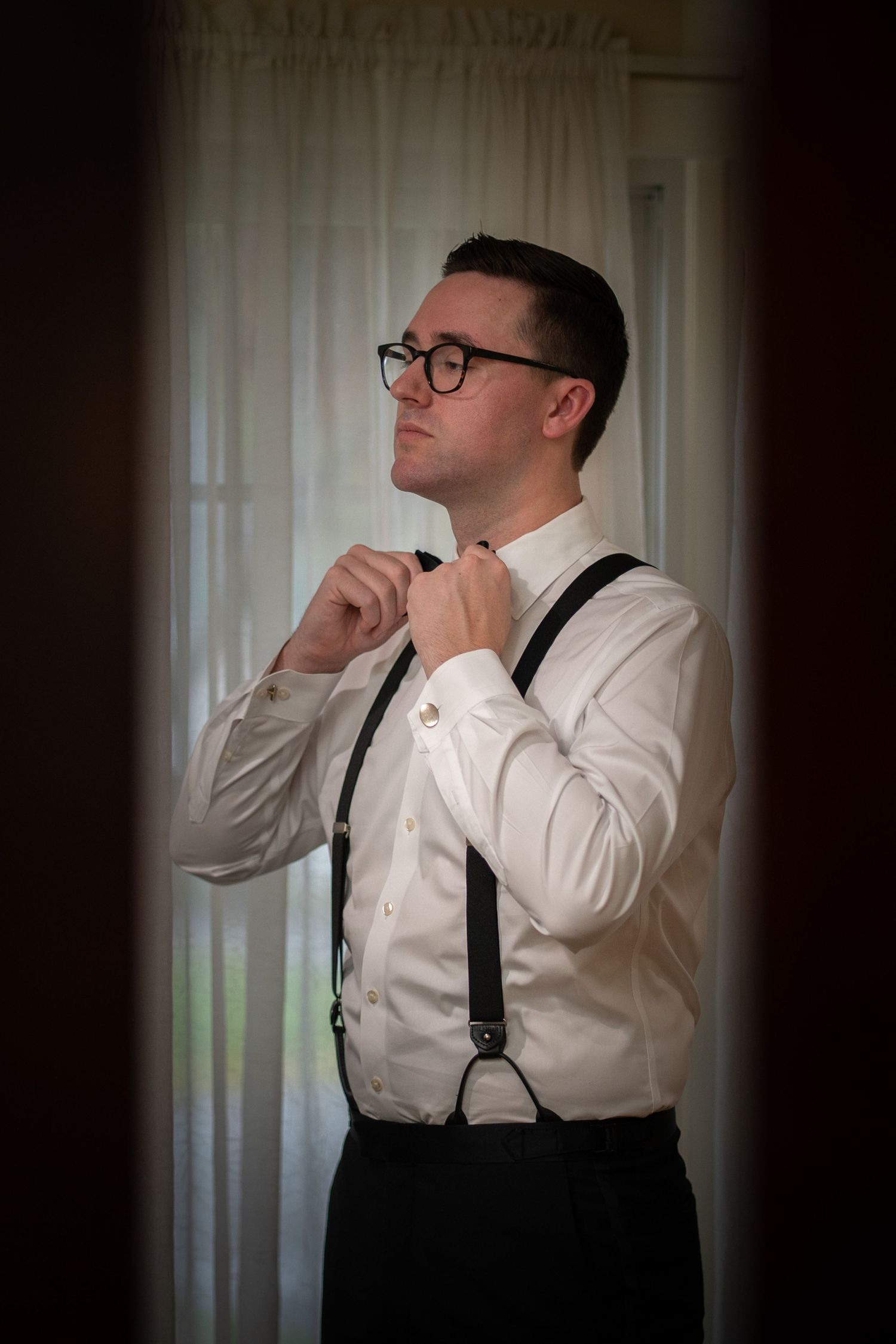 A groom in vintage-inspired attire adjusts his bowtie while wearing suspenders and glasses in a softly lit room.
