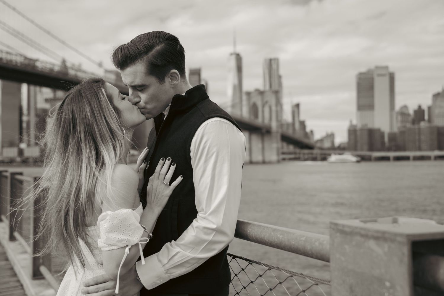 A romantic black and white photo of an affectionate couple by the Brooklyn Bridge with the Manhattan skyline in the background.