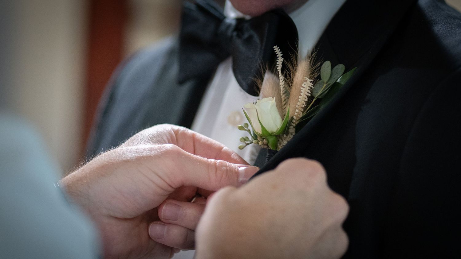 Someone adjusts a boutonniere with greenery and wheat details on a formal black tuxedo jacket.