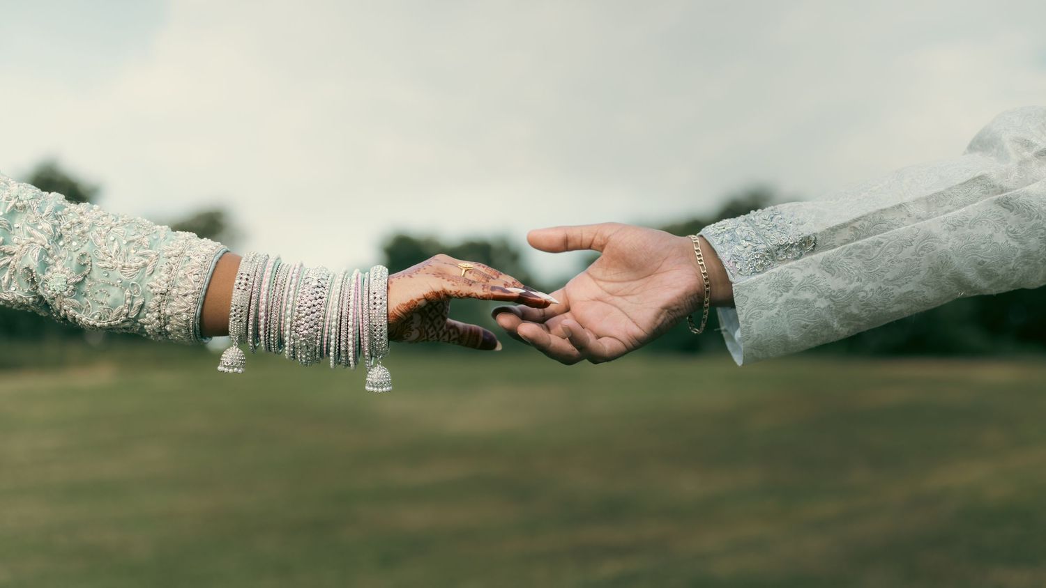 Two hands reaching toward each other with one wearing traditional Indian wedding bangles in a dreamy outdoor setting.