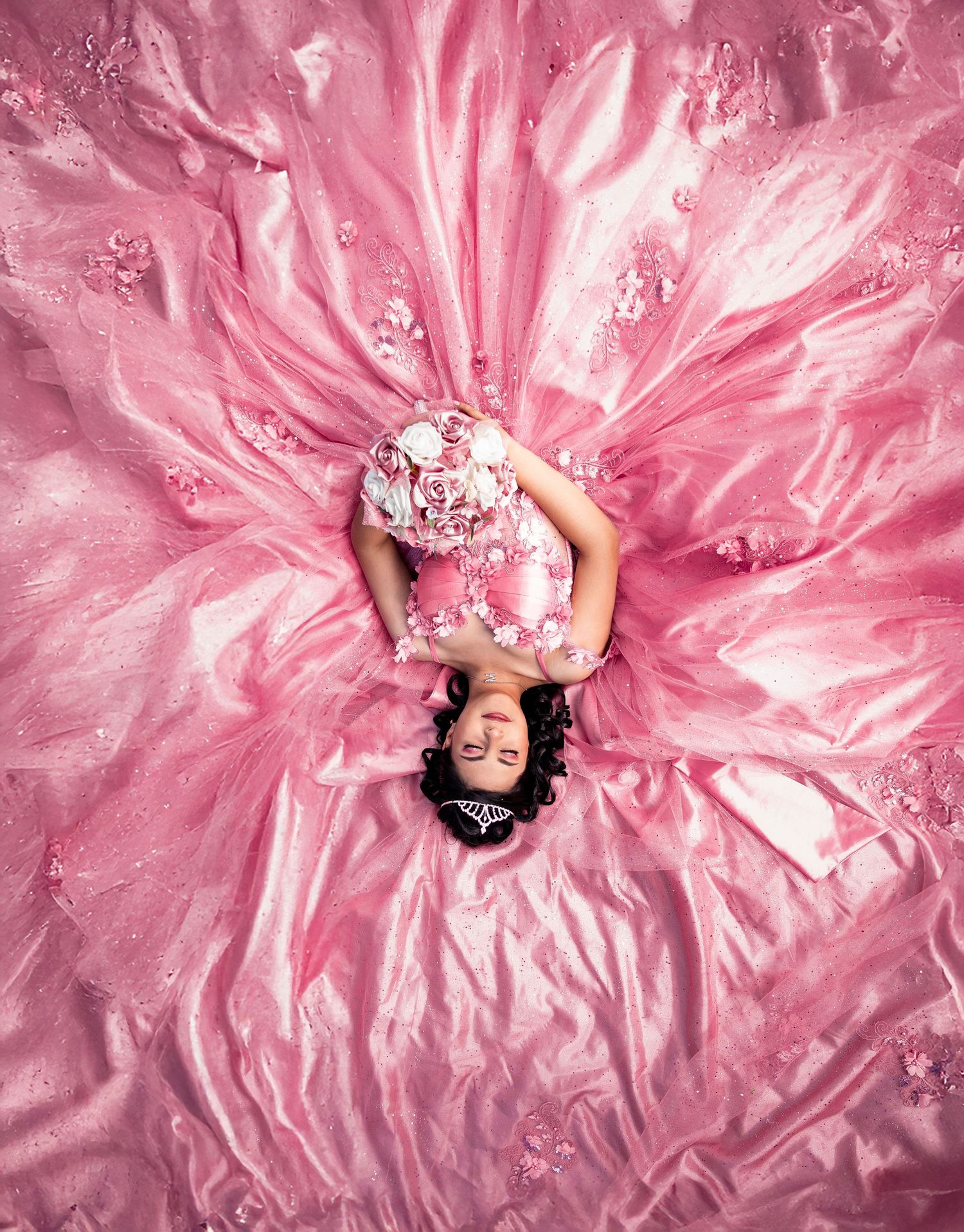 Overhead view of woman in floral dress lying on pink fabric creating dramatic skirt-like spread around her body.