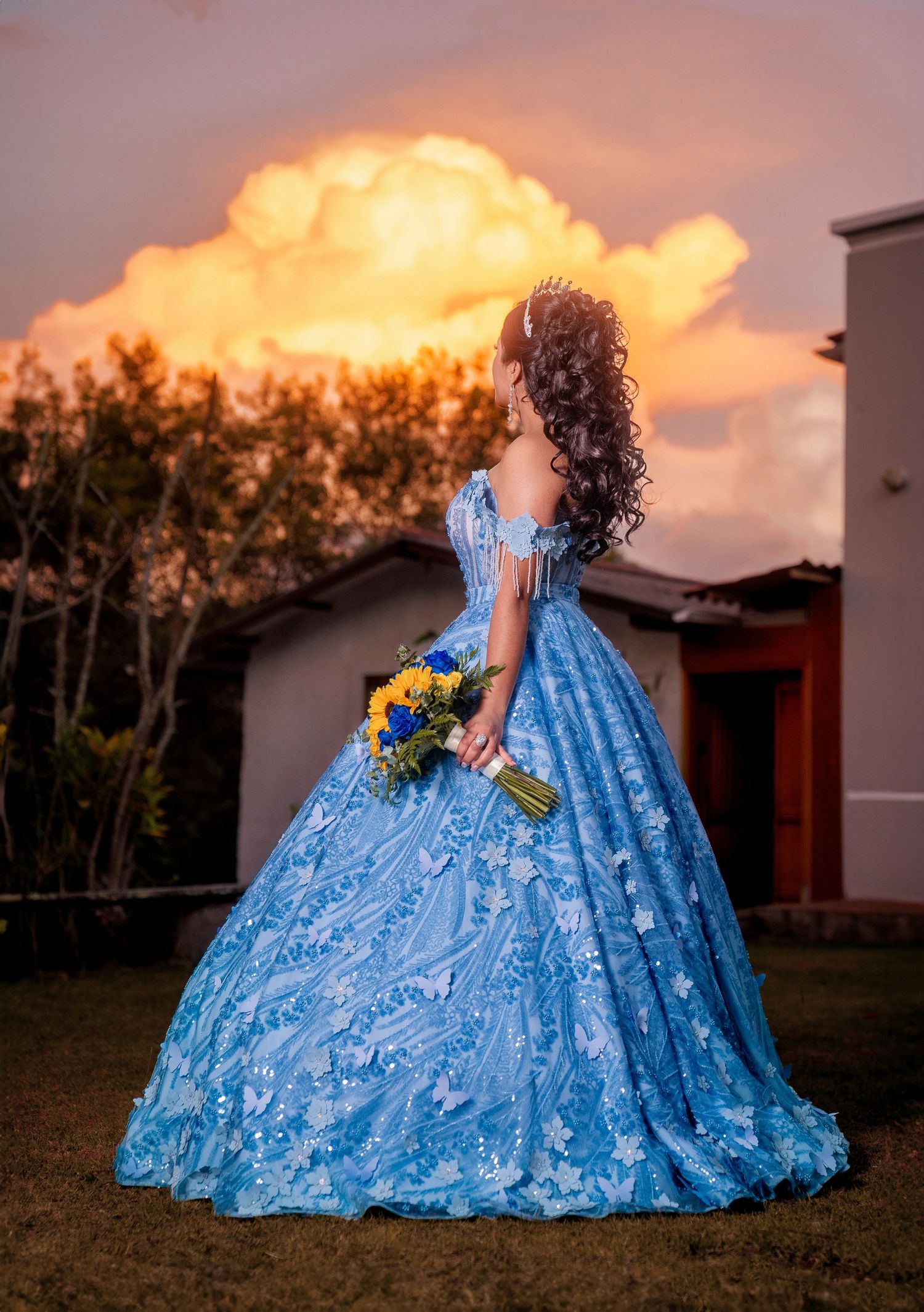 A person in a flowing light blue ballgown holds sunflowers while standing in a garden at sunset with dramatic clouds.