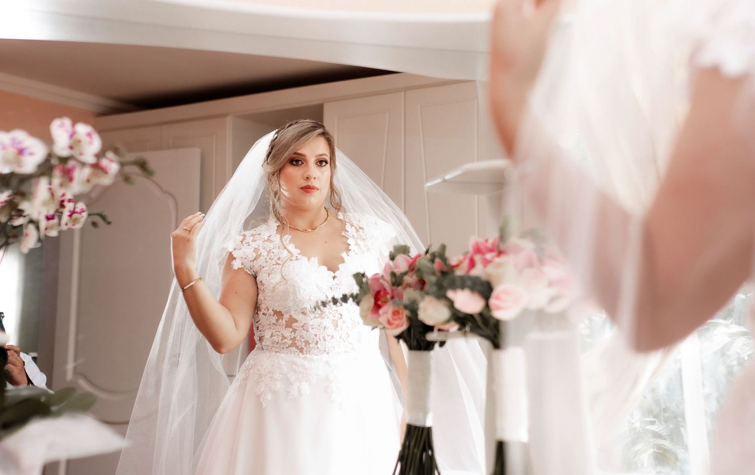 Bride in white lace gown looking at reflection in elegant room with pink floral decor.