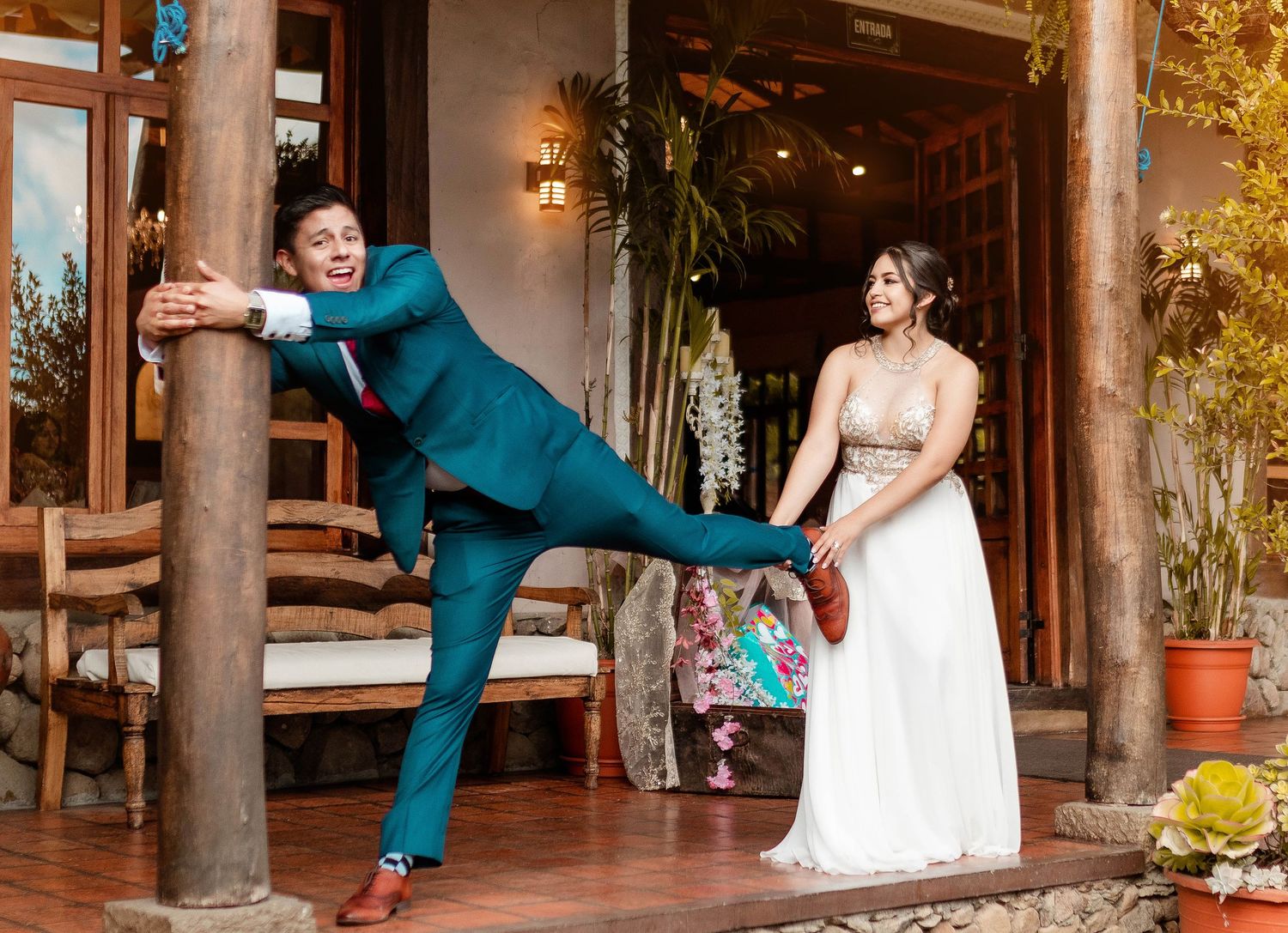 Playful wedding photo showing couple at rustic venue with groom striking dramatic pose against pillar.