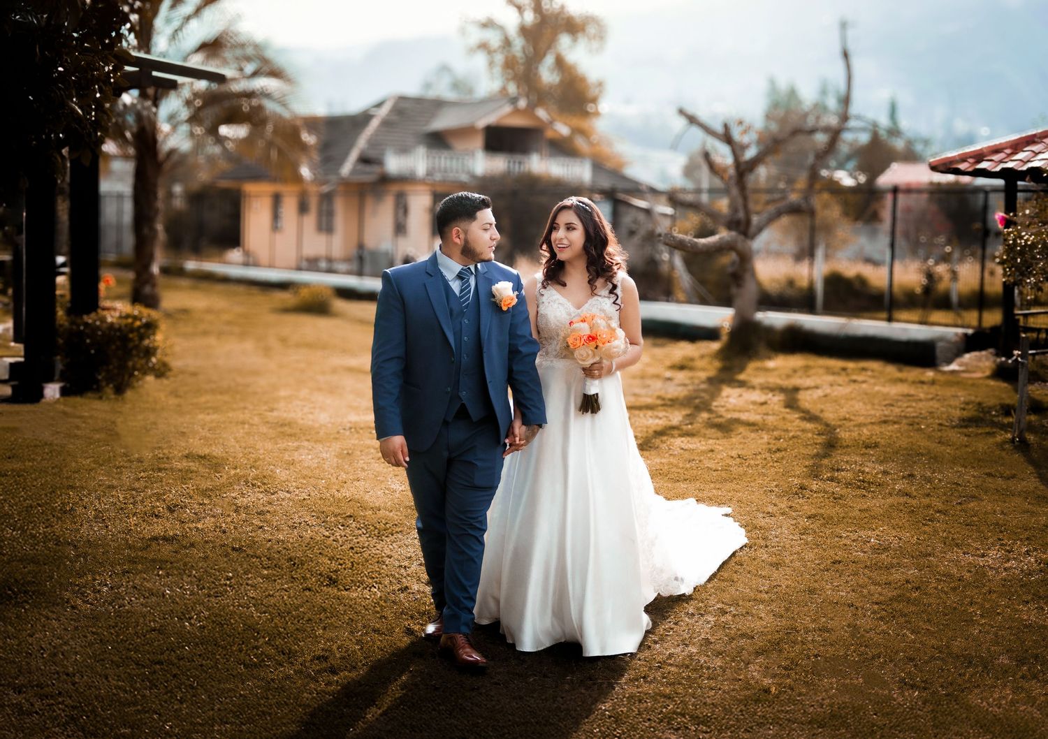 A couple in wedding attire walks together on a rustic autumn path with a white house in the background.