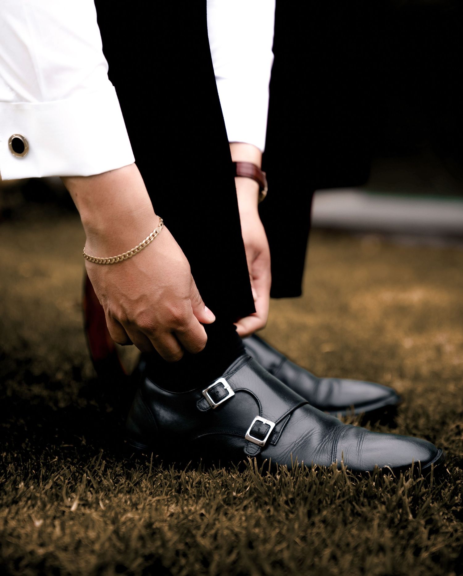 Person wearing a white dress shirt and black pants adjusts a pair of polished double monk strap dress shoes on grass.