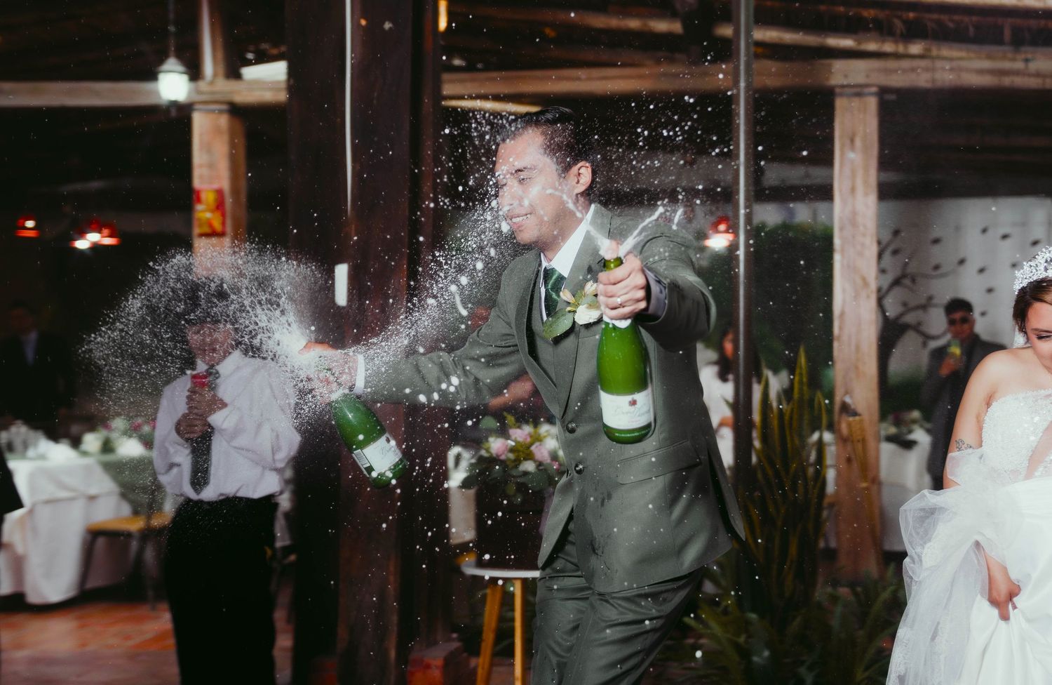 Champagne bottle sprays celebratory bubbles during a festive wedding reception moment in an indoor venue.