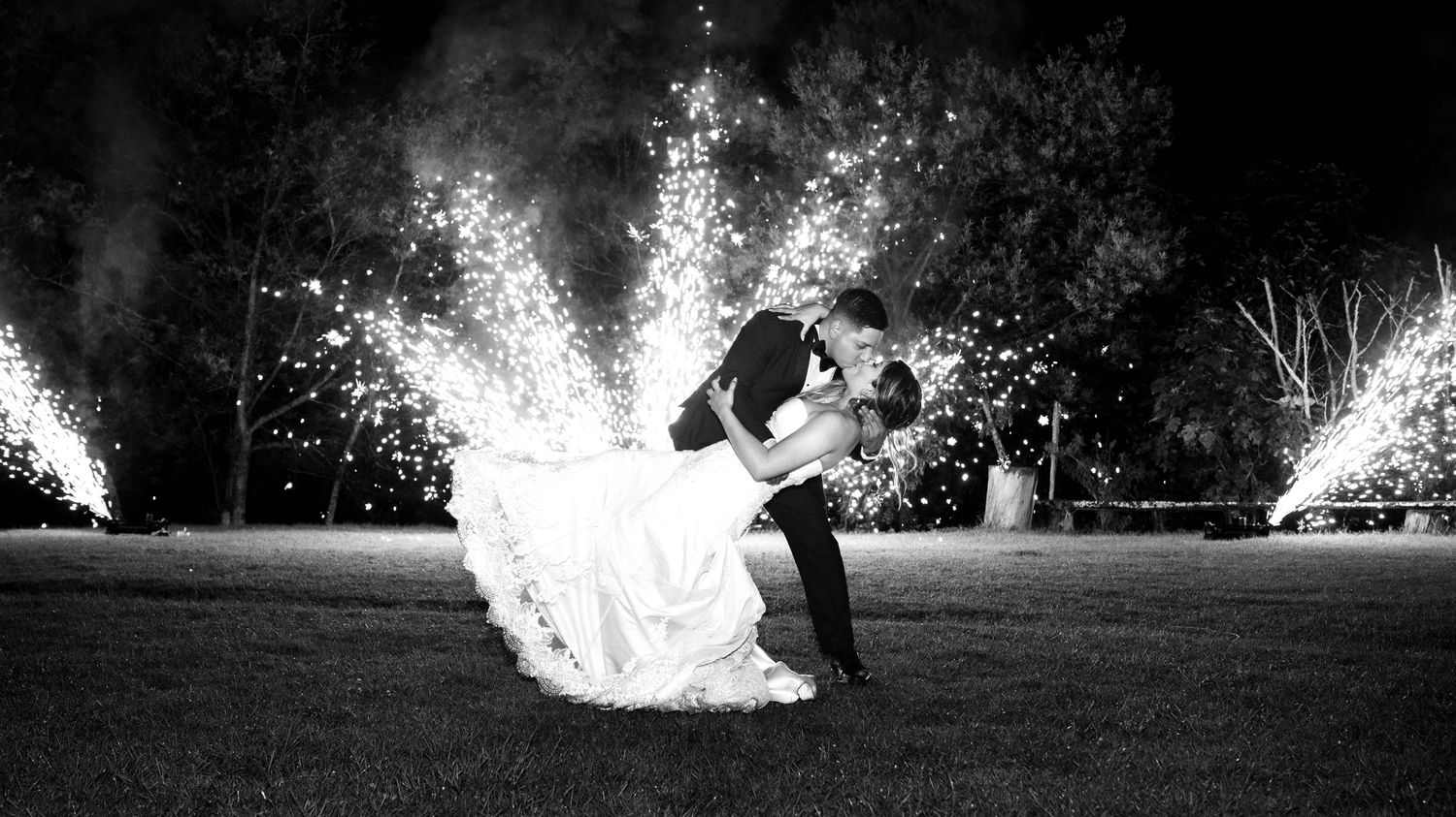 A romantic couple embraces in black and white while sparklers create dramatic light effects around them on a grassy lawn.