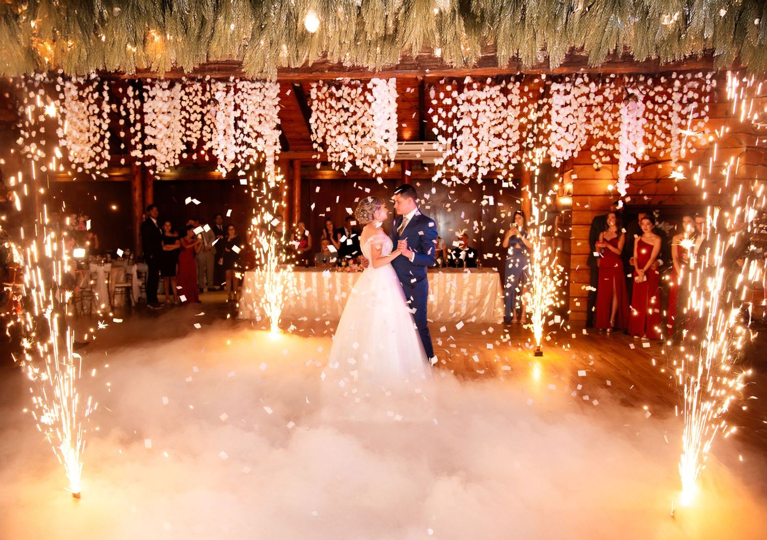 Couple shares their first dance at wedding reception under glowing string lights and sparkling pyrotechnic effects.