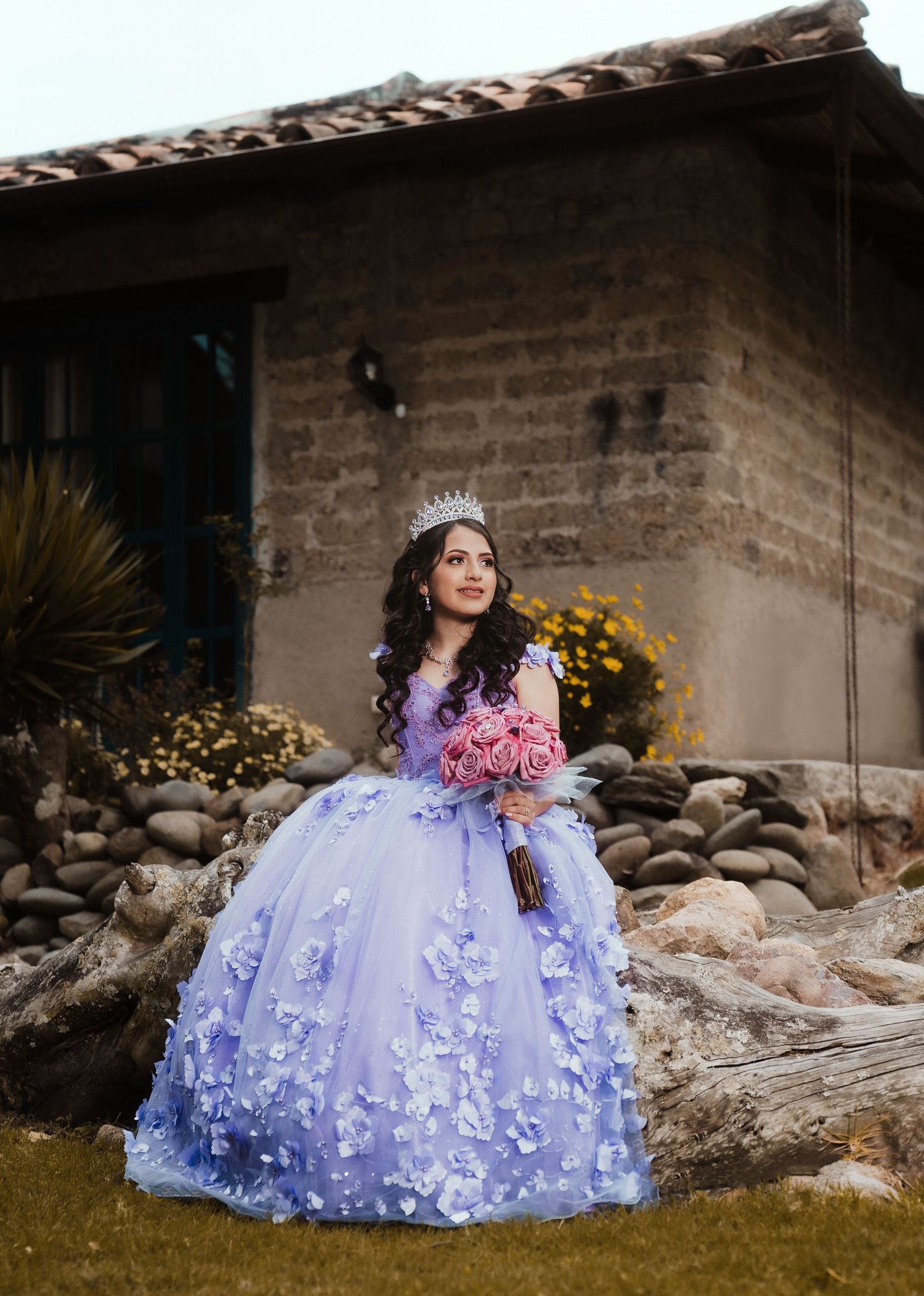 A person in a lavender quinceañera ball gown with floral appliques poses outdoors by a rustic stone wall.