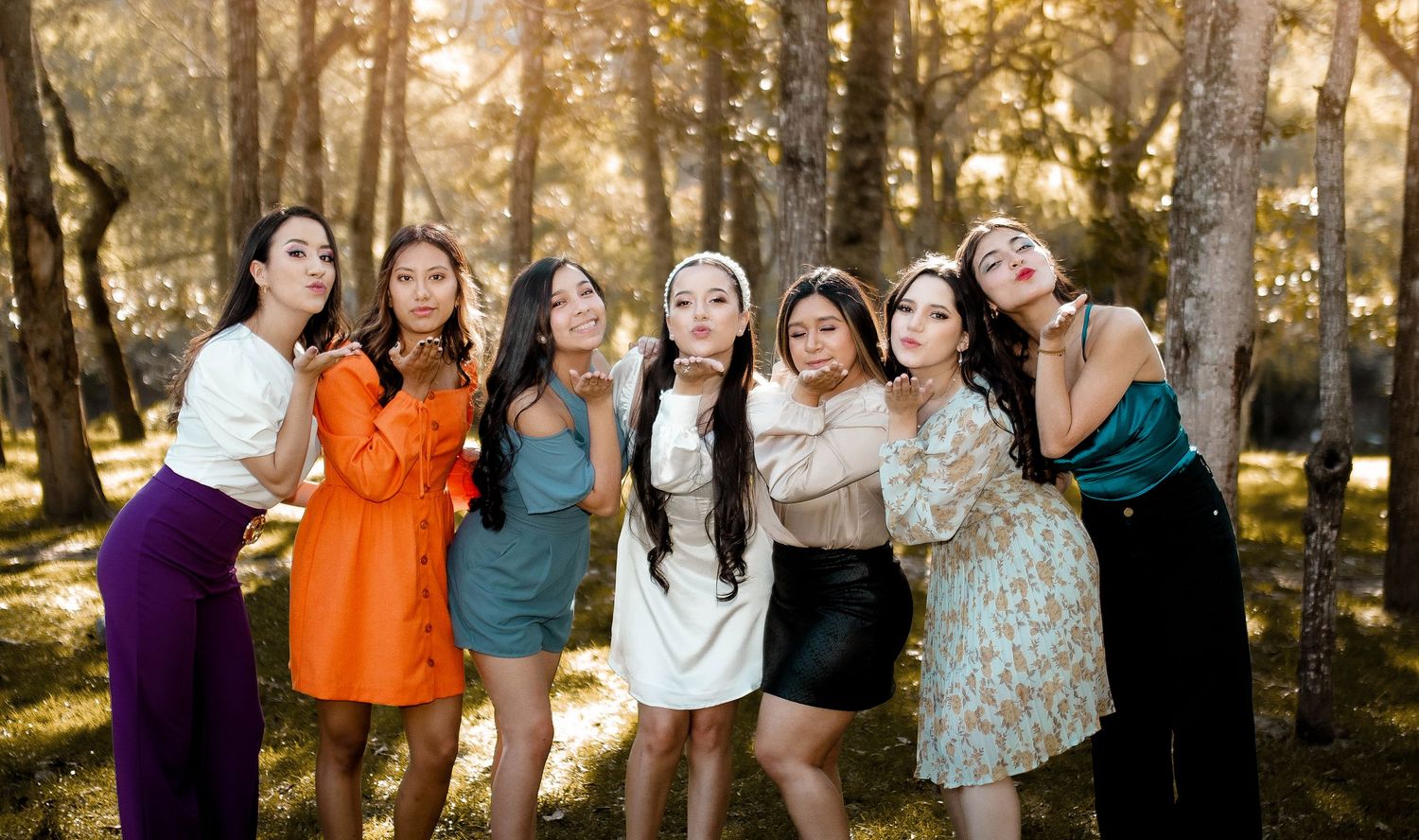 Group of friends in colorful dresses pose together in sunlit forest setting.
