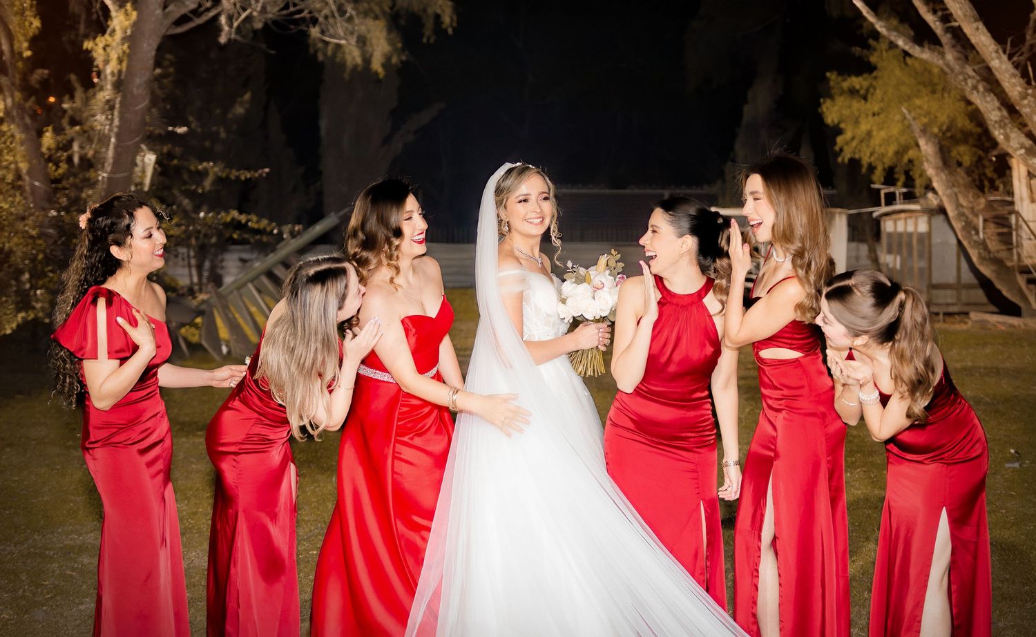 Wedding party in red bridesmaid dresses sharing candid moment outdoors at night.