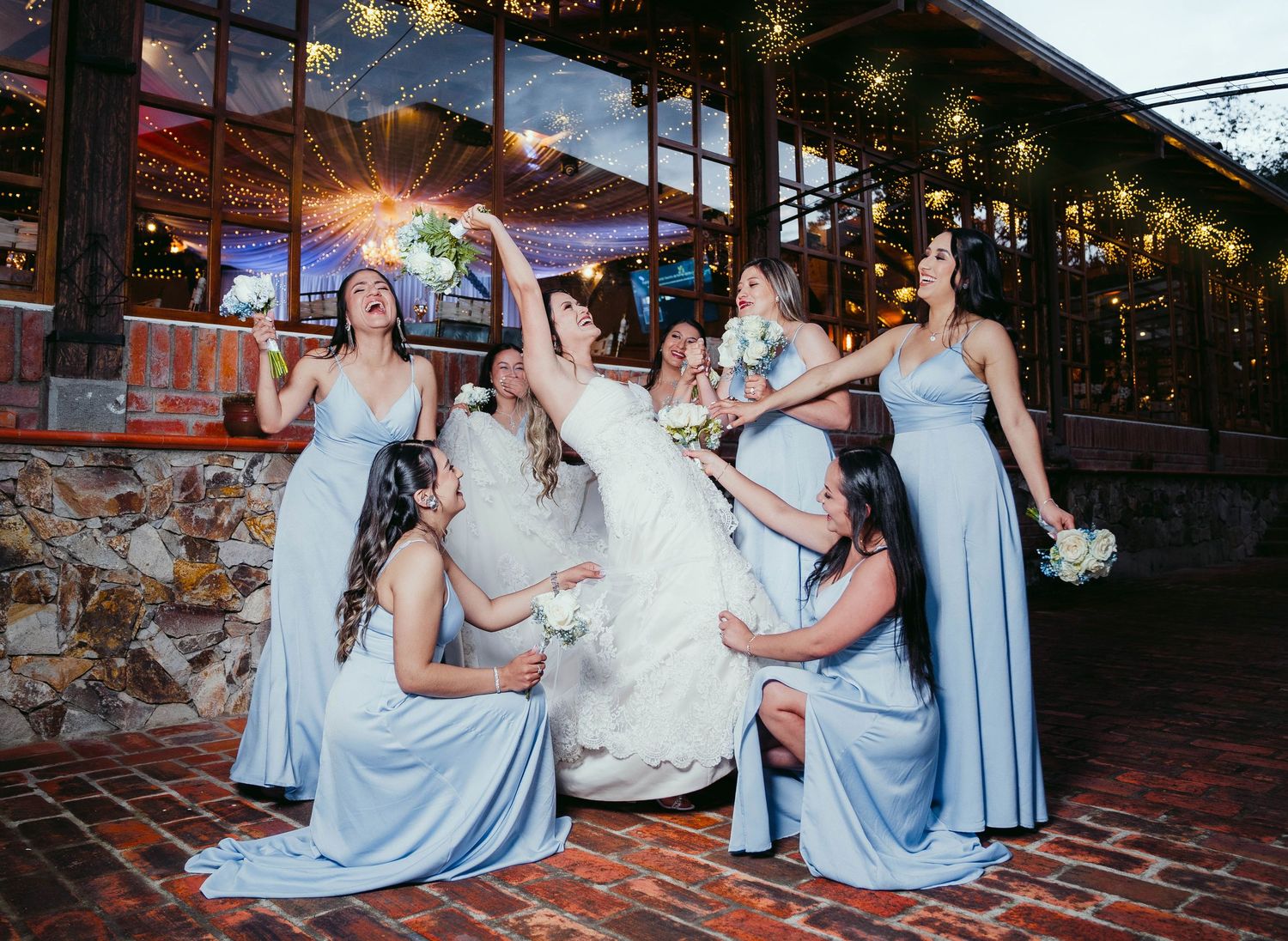 A wedding party poses on brick steps at dusk with string lights glowing above in an outdoor garden setting.
