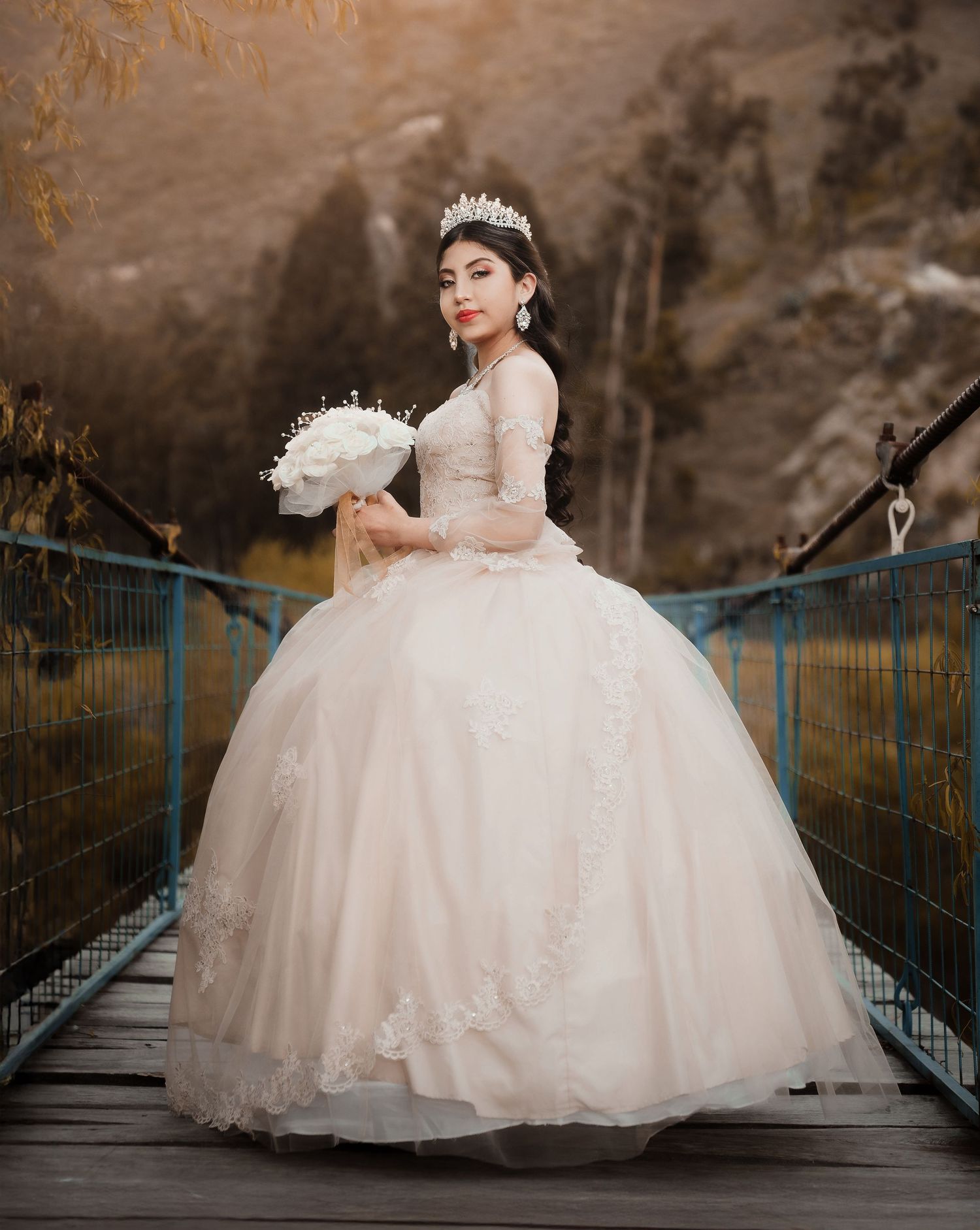 Bride in elegant white ball gown and tiara poses on rustic bridge during autumn outdoor photoshoot.