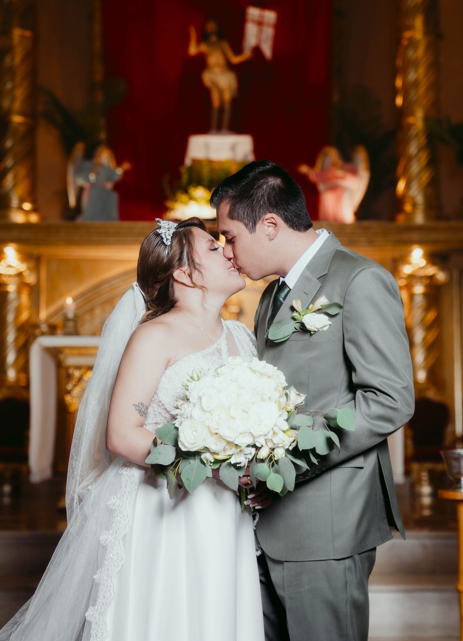 A couple shares their first kiss as newlyweds at the altar in a church with white bouquet and grey suit.