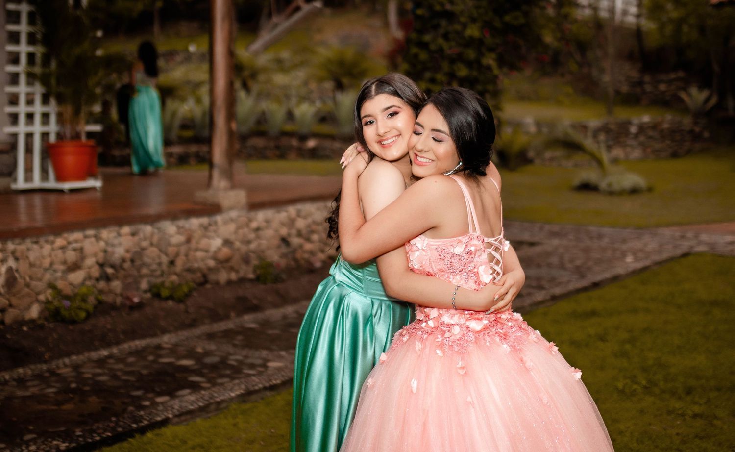 Two people embracing while wearing formal gowns in garden setting.