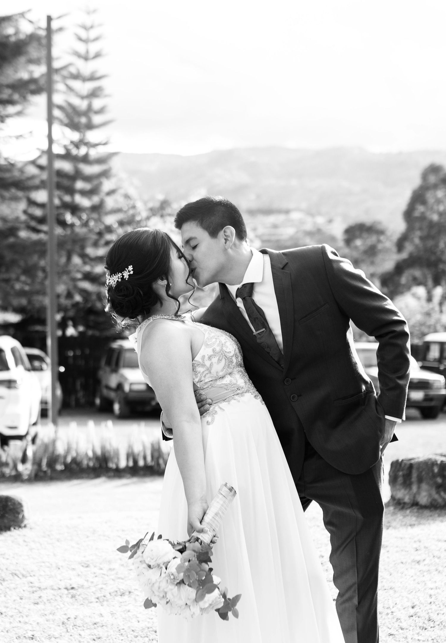 A romantic black and white wedding photo of a couple sharing a kiss outdoors with mountains in the background.