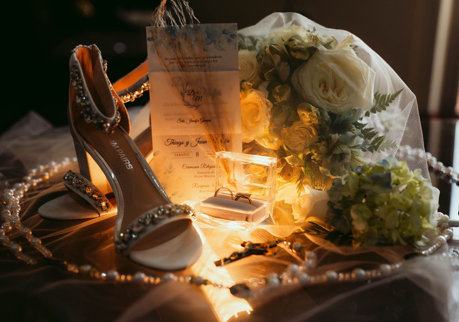 Burning candles cast a warm golden glow among wedding details and scattered white flowers on a dark surface.