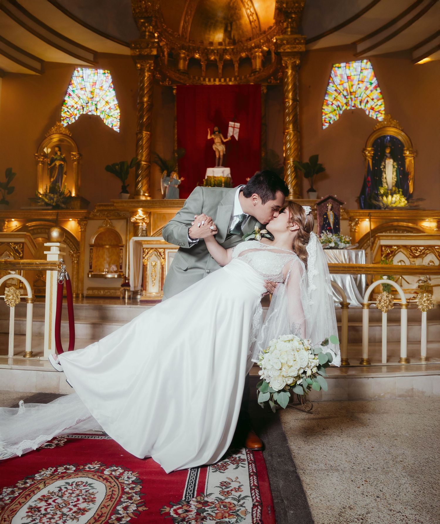 Couple shares a romantic dip kiss at the altar in an ornate church interior with stained glass windows and gold decor.
