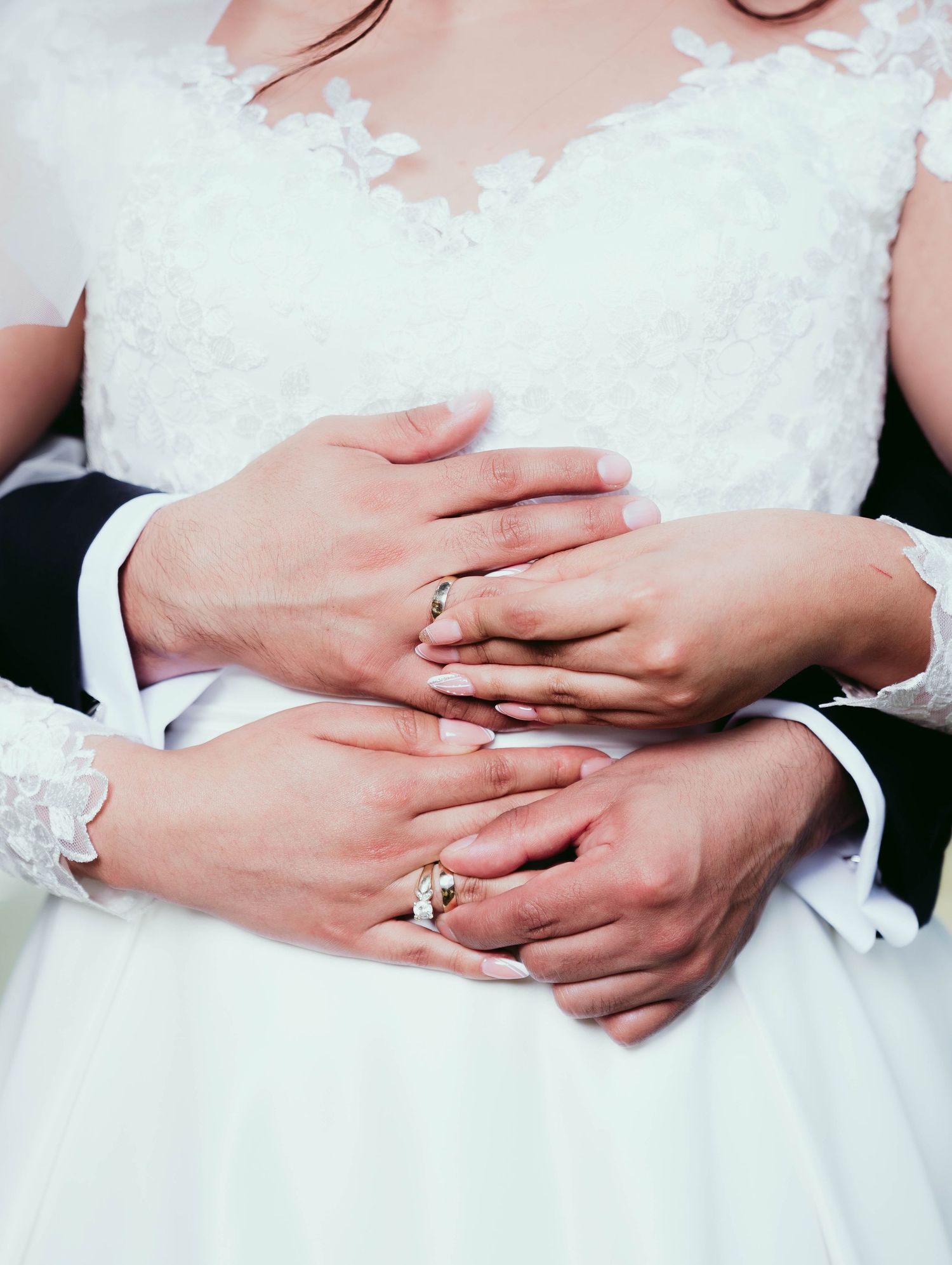 Newlywed couple's hands with wedding rings embrace around bride's white lace dress waist.