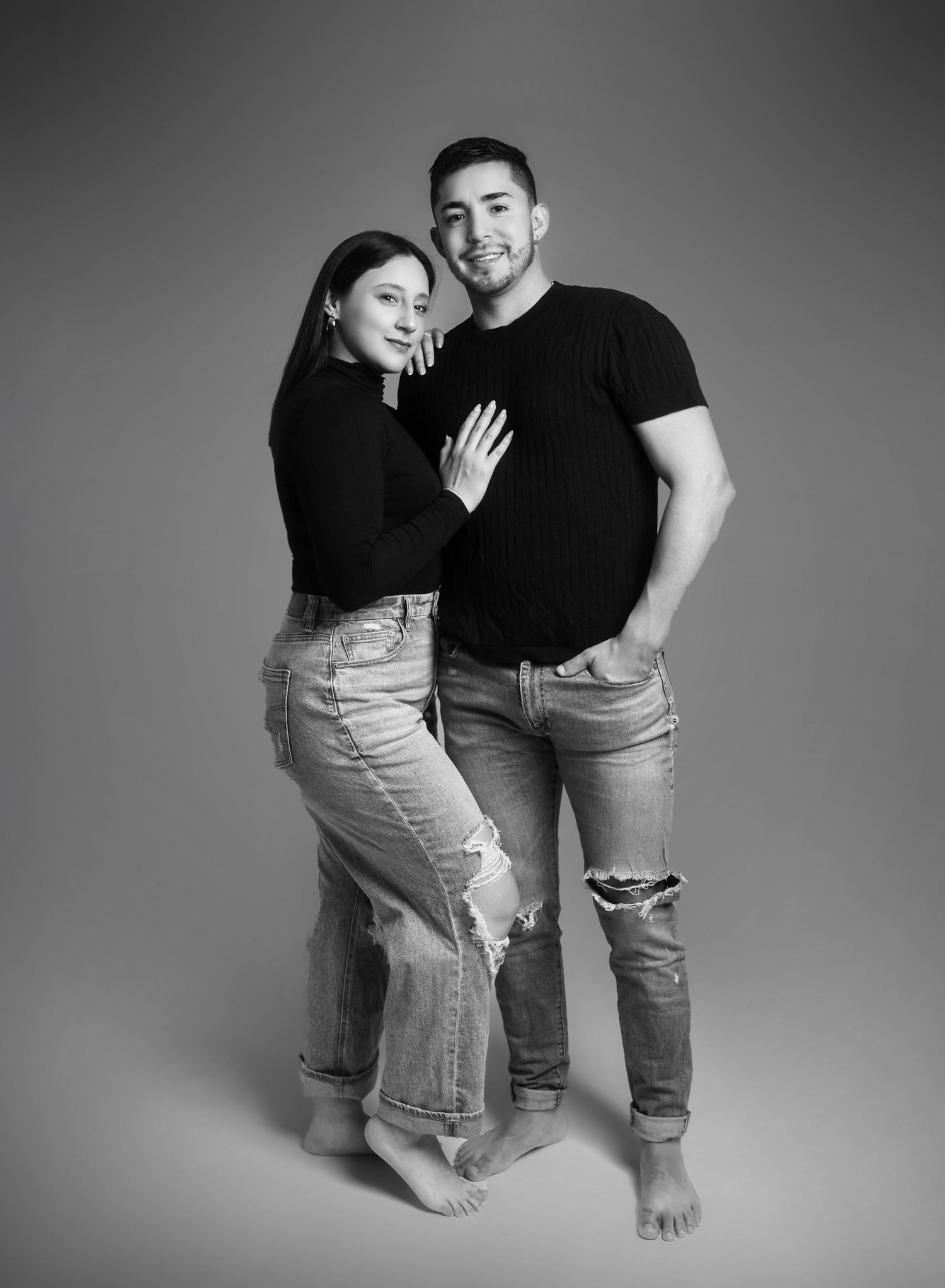 Black and white studio portrait of couple in casual wear against gray background.