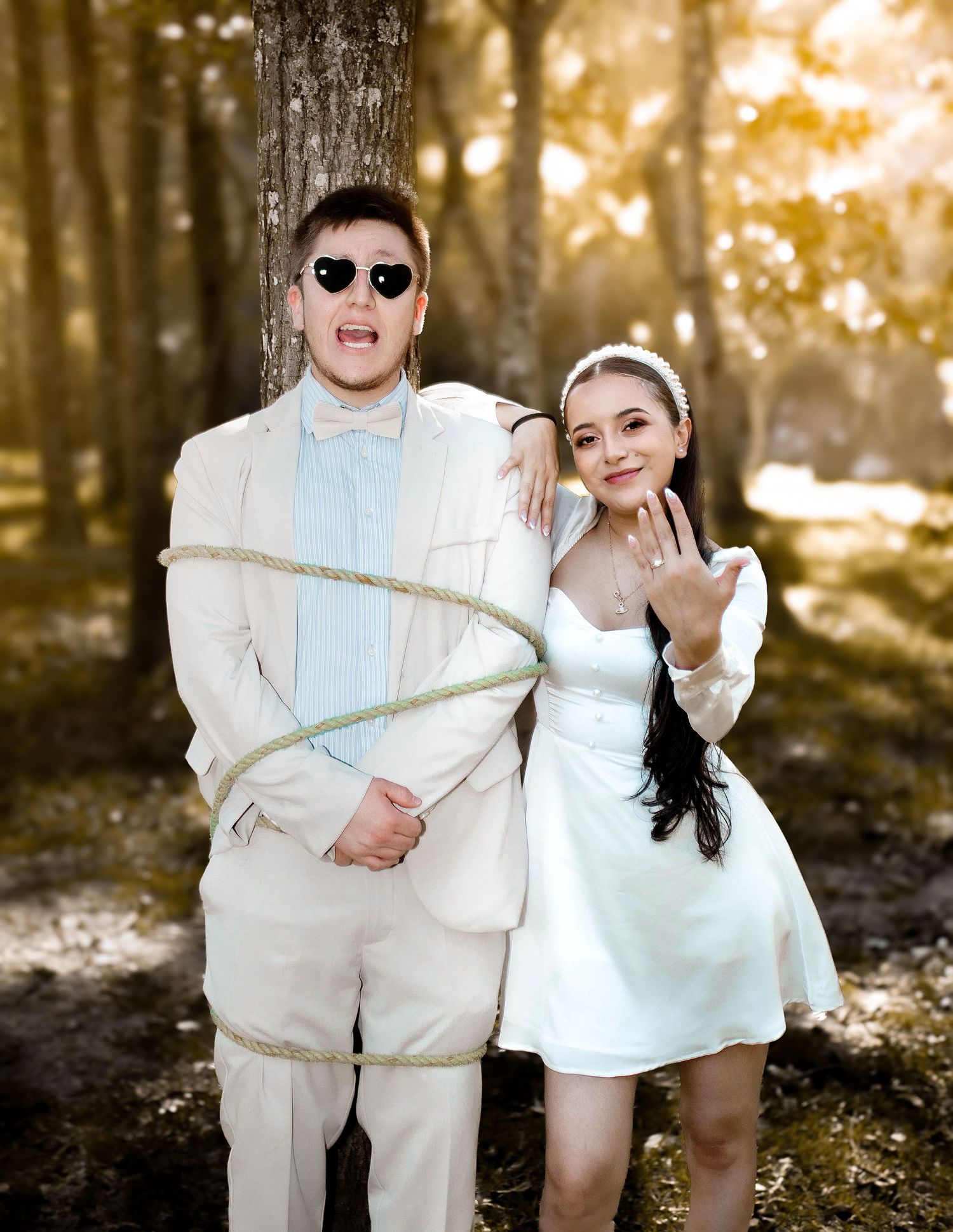 A playful wedding photo with the groom tied up while bride shows off her ring in a forest setting.