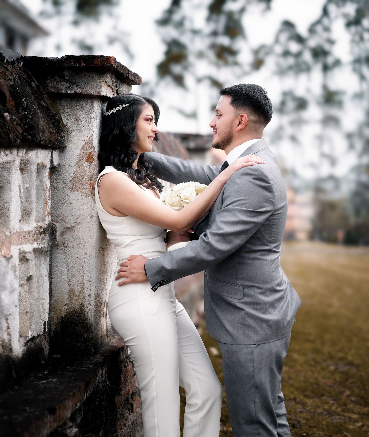 A happy couple embraces in a romantic outdoor setting with a rustic stone wall backdrop and soft dreamy lighting.
