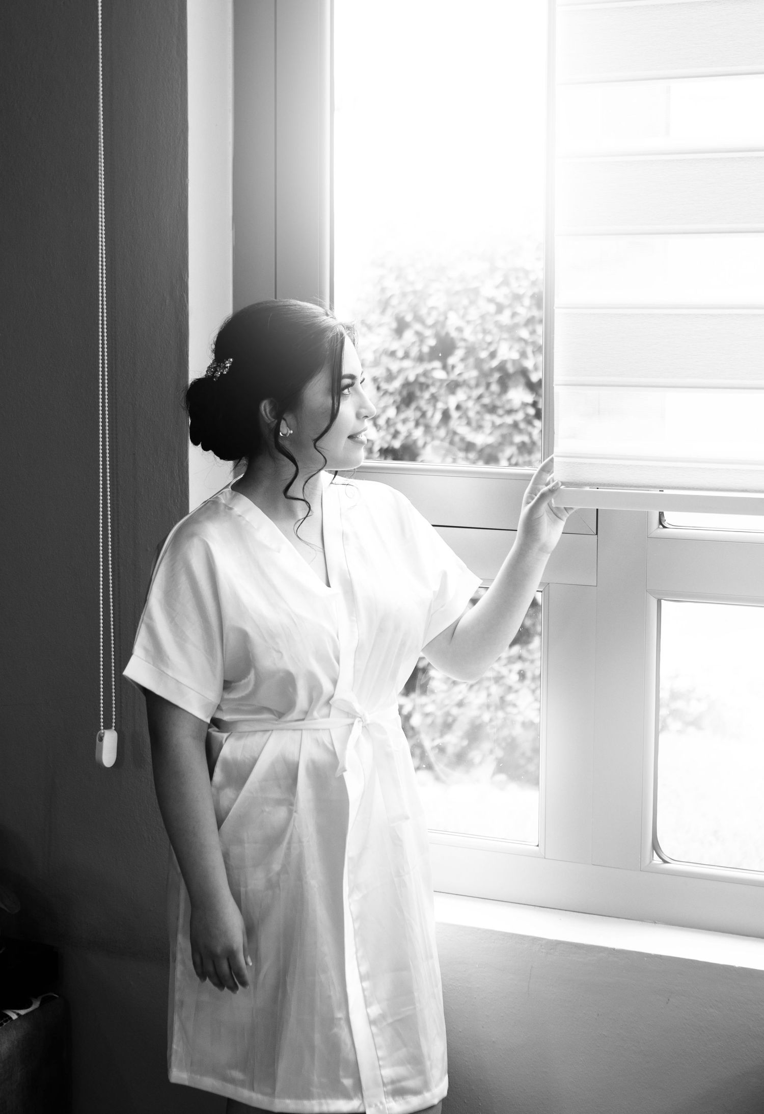 Figure in white robe gazing through window with soft natural lighting in black and white.